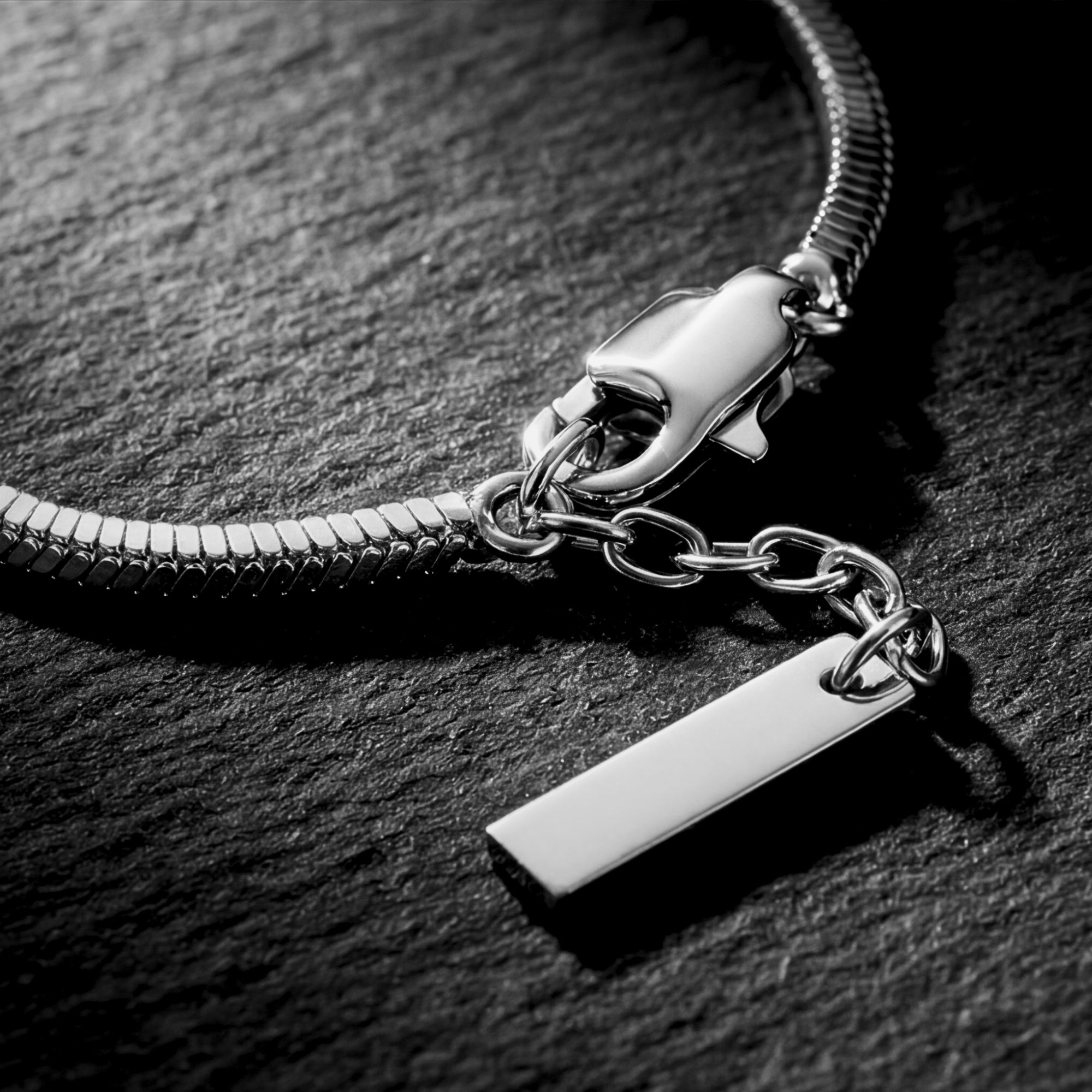 Macro detail of the lobster clasp and rectangular tag on a silver stainless steel square link bracelet.