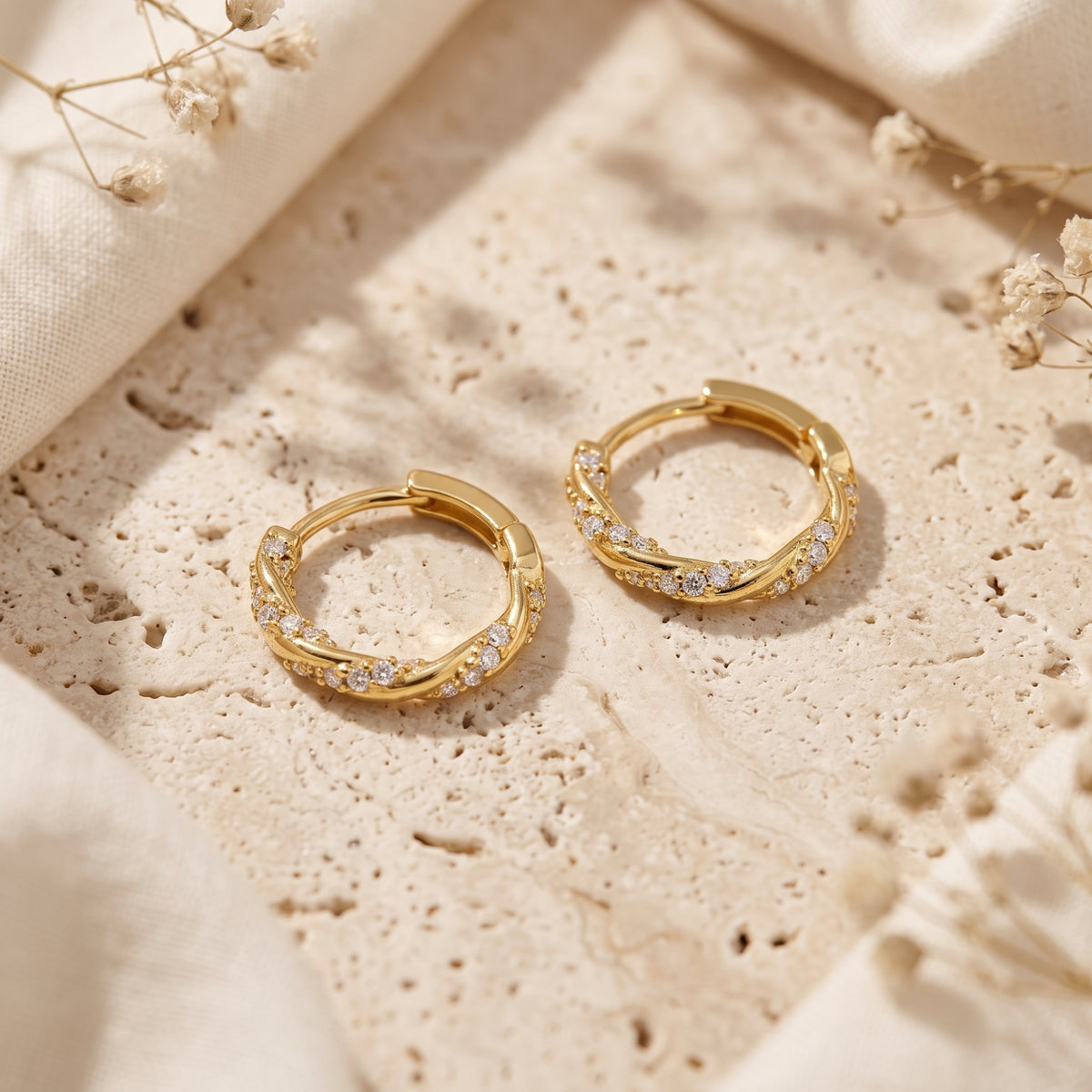 Two gold-tone twisted hoop earrings with clear cubic zirconia placed on textured stone among fabric and dried flowers.