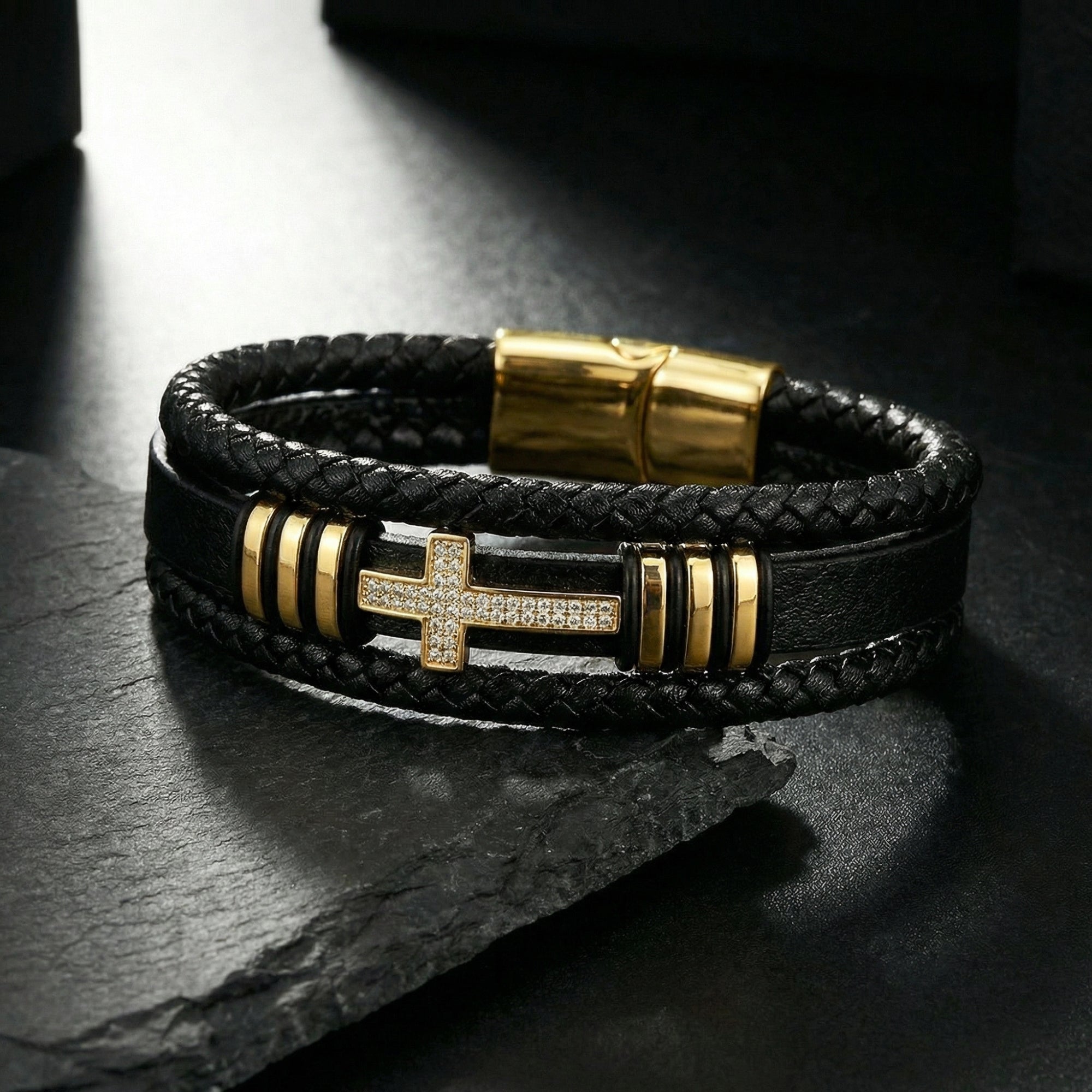 Close-up of a gold-finished stainless steel cross with pavé stones on a multi-layered black leather bracelet.