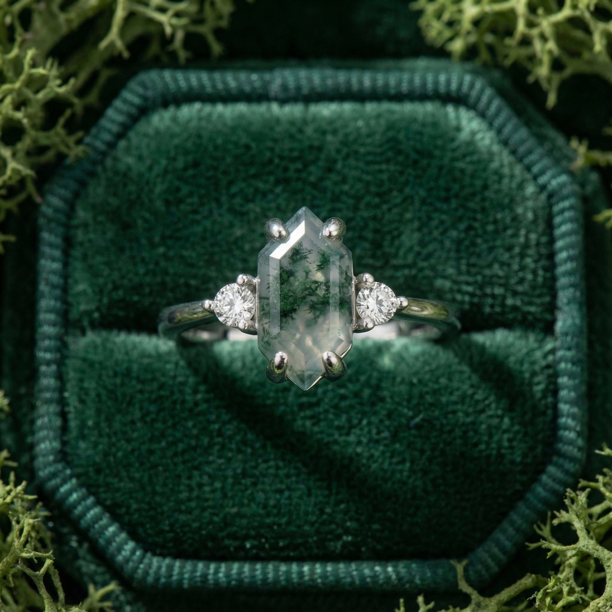 Macro view of a hexagon cut moss agate ring with round side stones in a green velvet jewelry box.