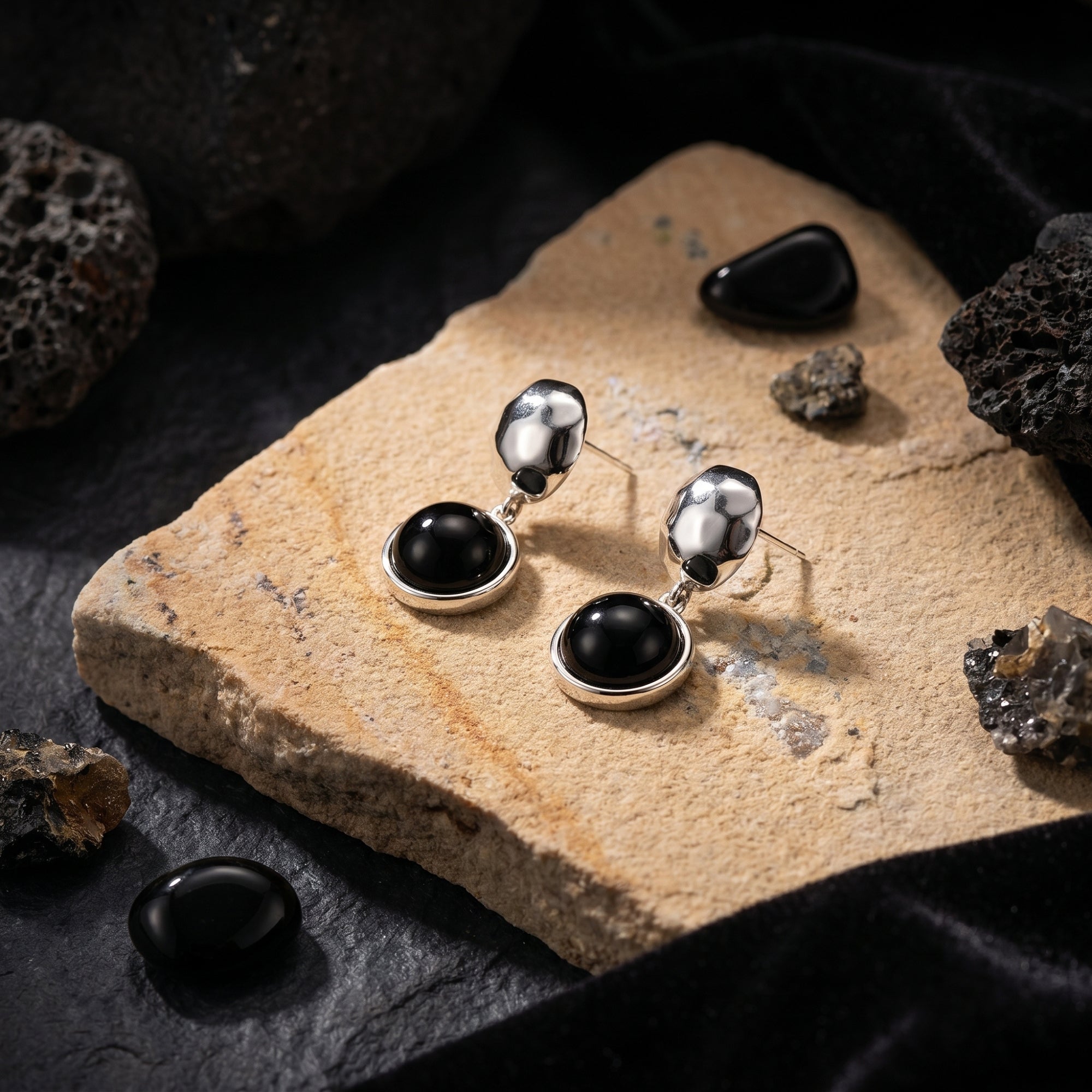 Angled view of silver hammered drop earrings with round black onyx stones resting on a textured tan stone.