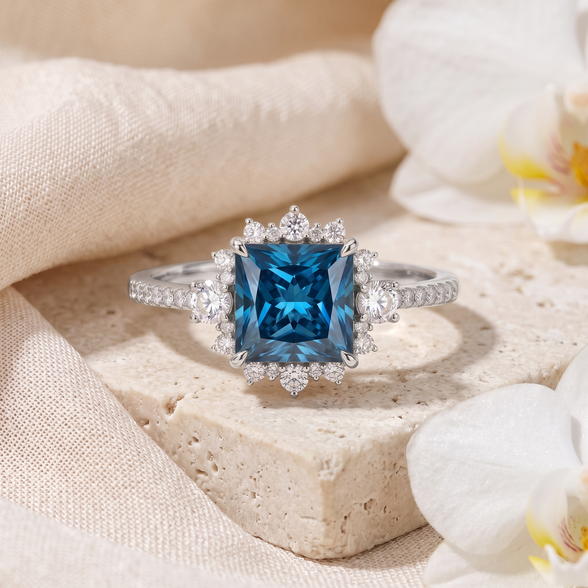 Princess-cut blue moissanite halo engagement ring in white gold resting on a stone slab with floral accents.