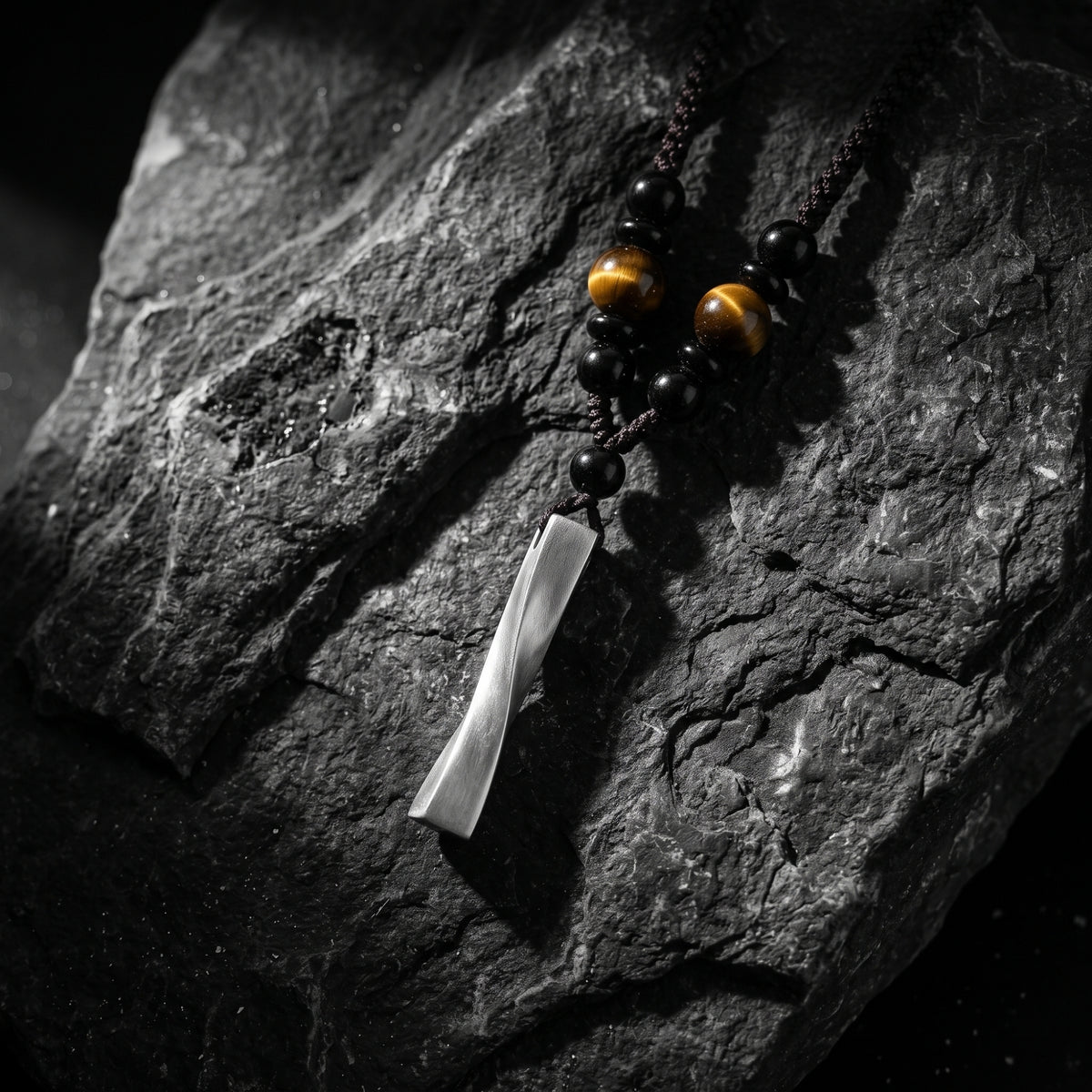 Twisted silver bar pendant resting diagonally with black and tiger eye beads on a dark textured rock.