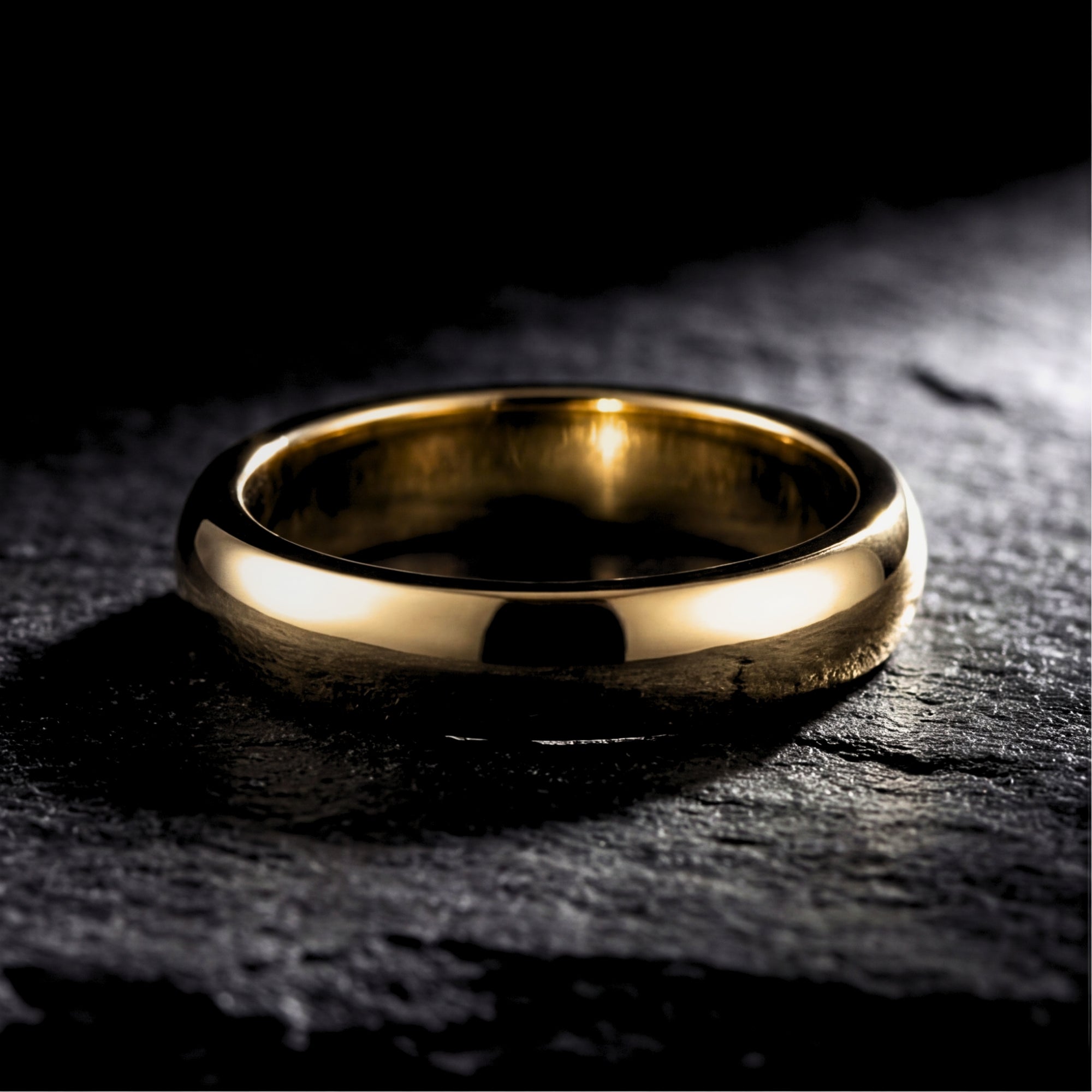 Close-up of a gold tone stainless steel band ring lying on a dark, textured stone surface.