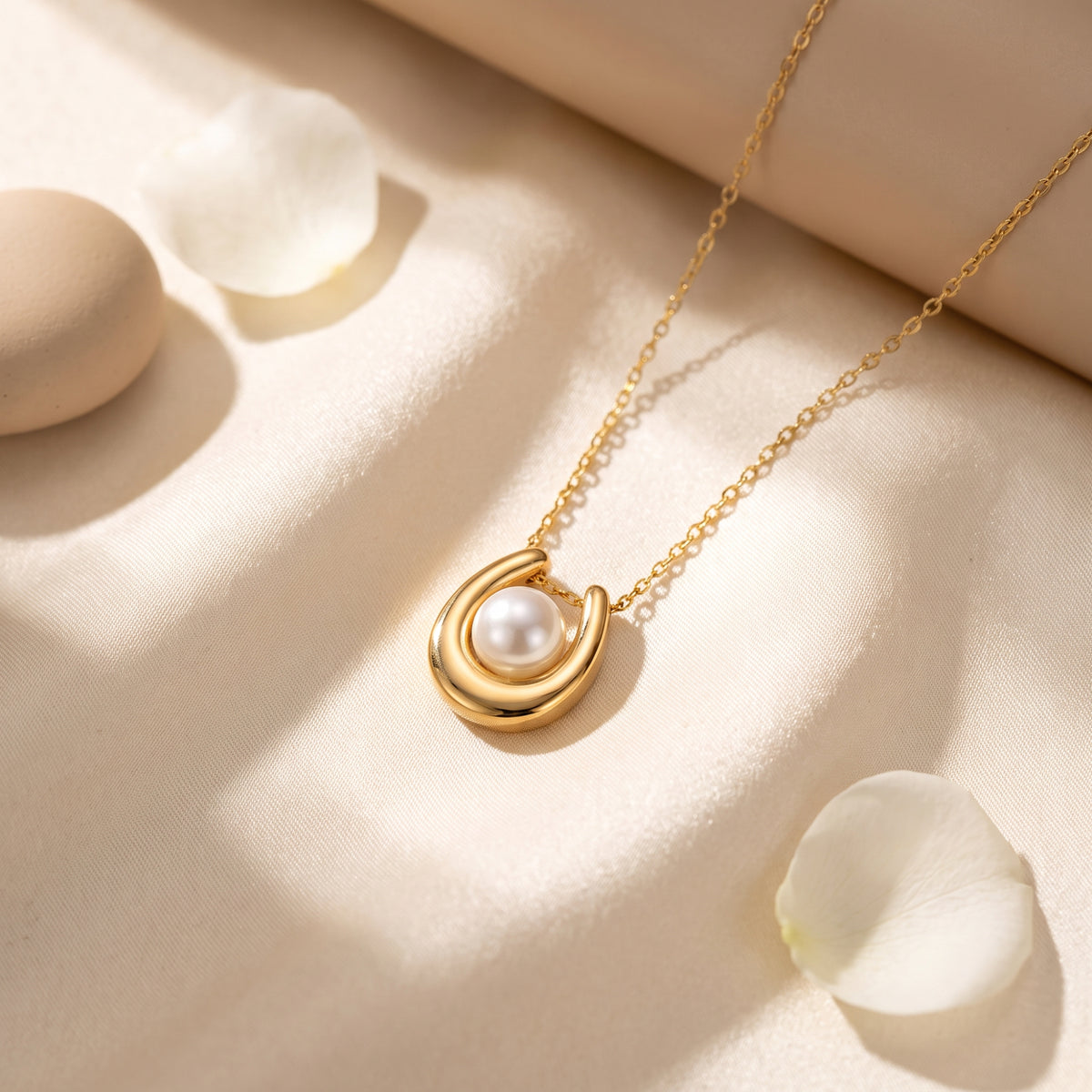 Gold-tone horseshoe pearl pendant resting flat on cream fabric beside white flower petals.