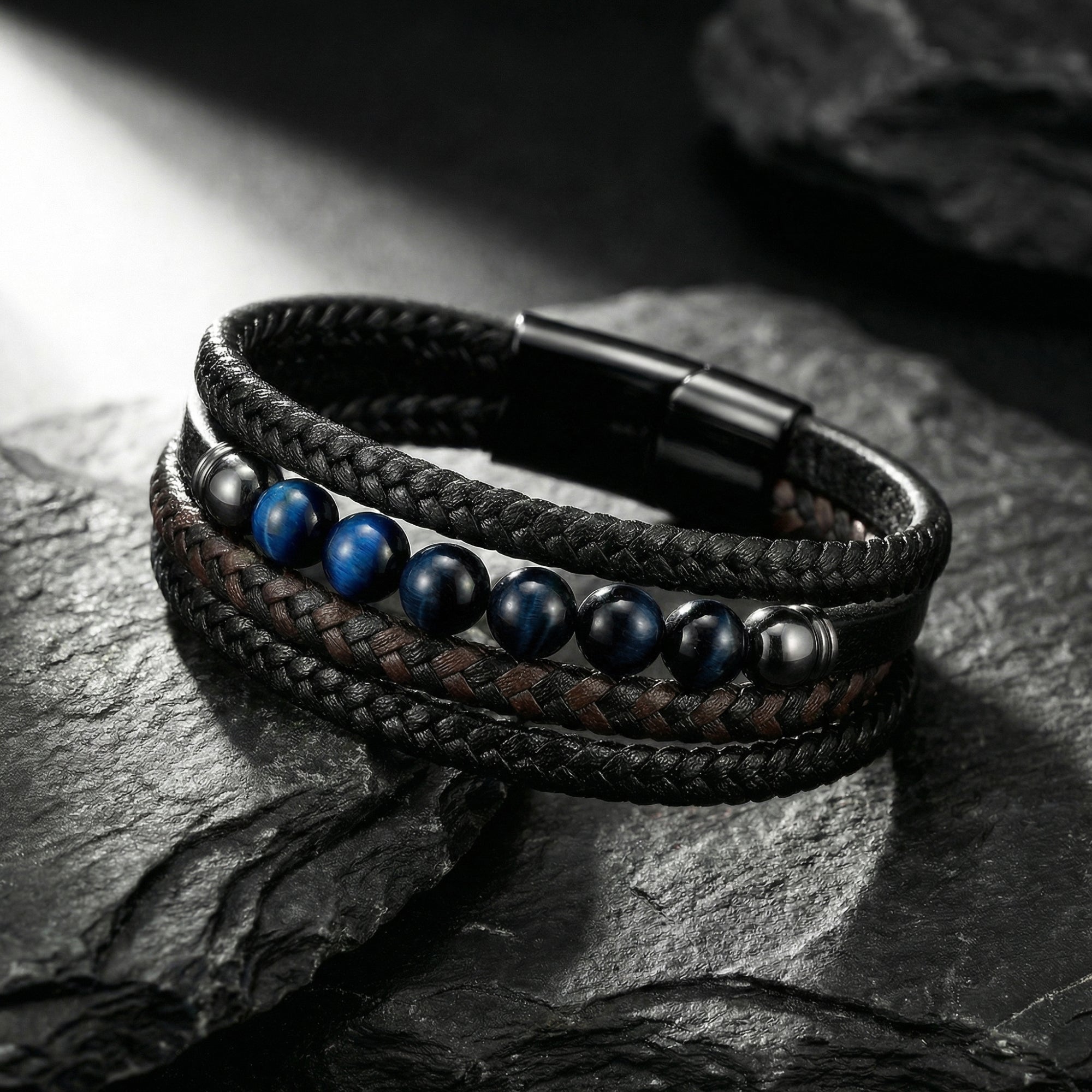 Braided black and brown leather bracelet with blue accent beads and black steel clasp on a dark stone background.