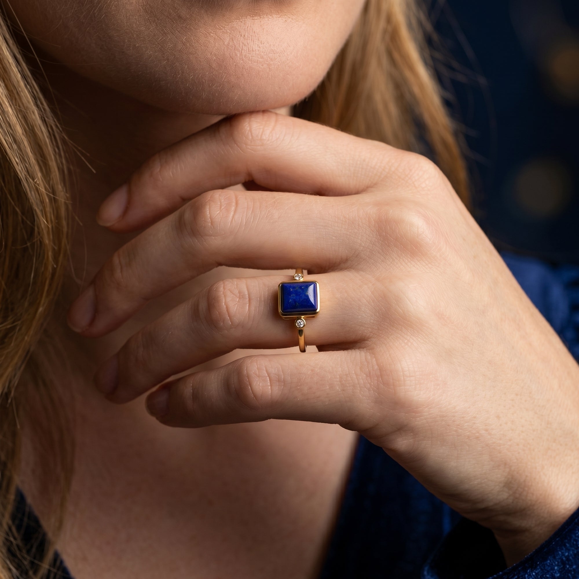 Close-up of a woman's hand wearing a yellow gold ring with a rectangular natural lapis lazuli gemstone.
