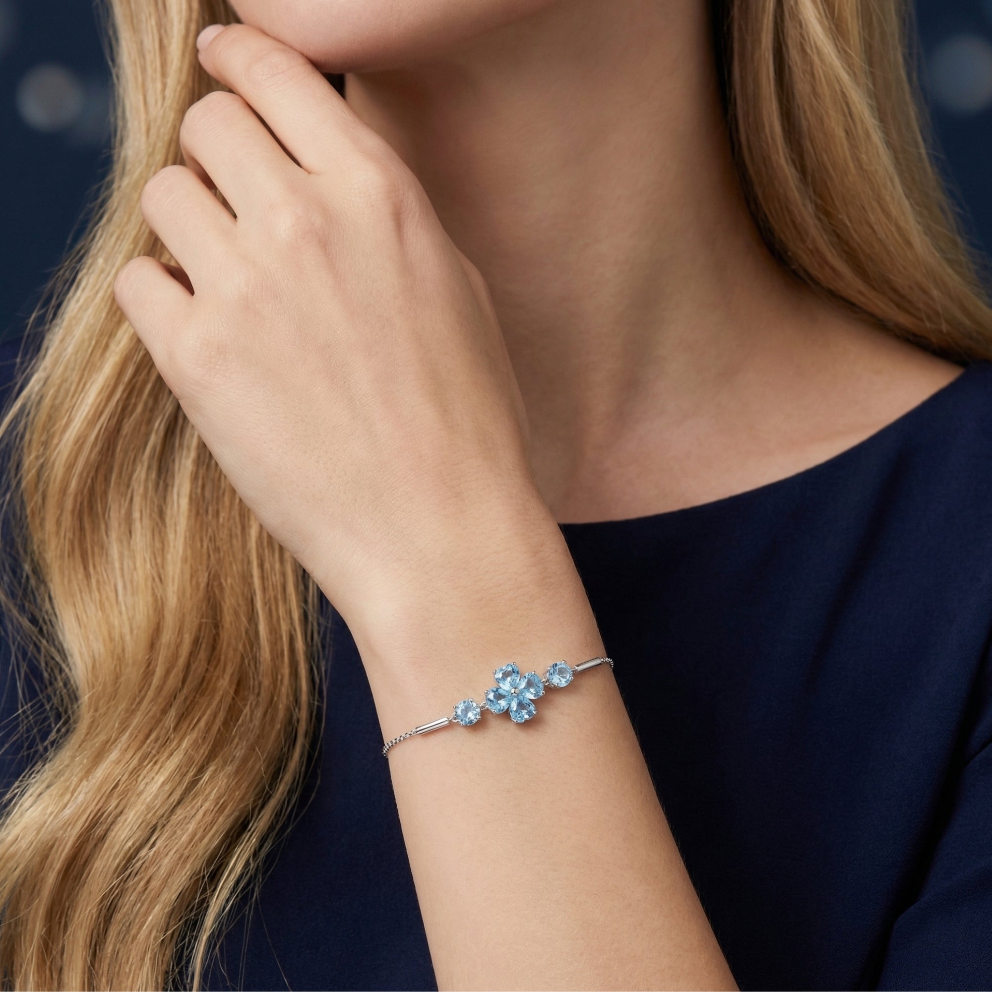 Silver Swiss blue topaz clover bracelet with heart and round cuts worn on a woman's wrist against a blue dress.