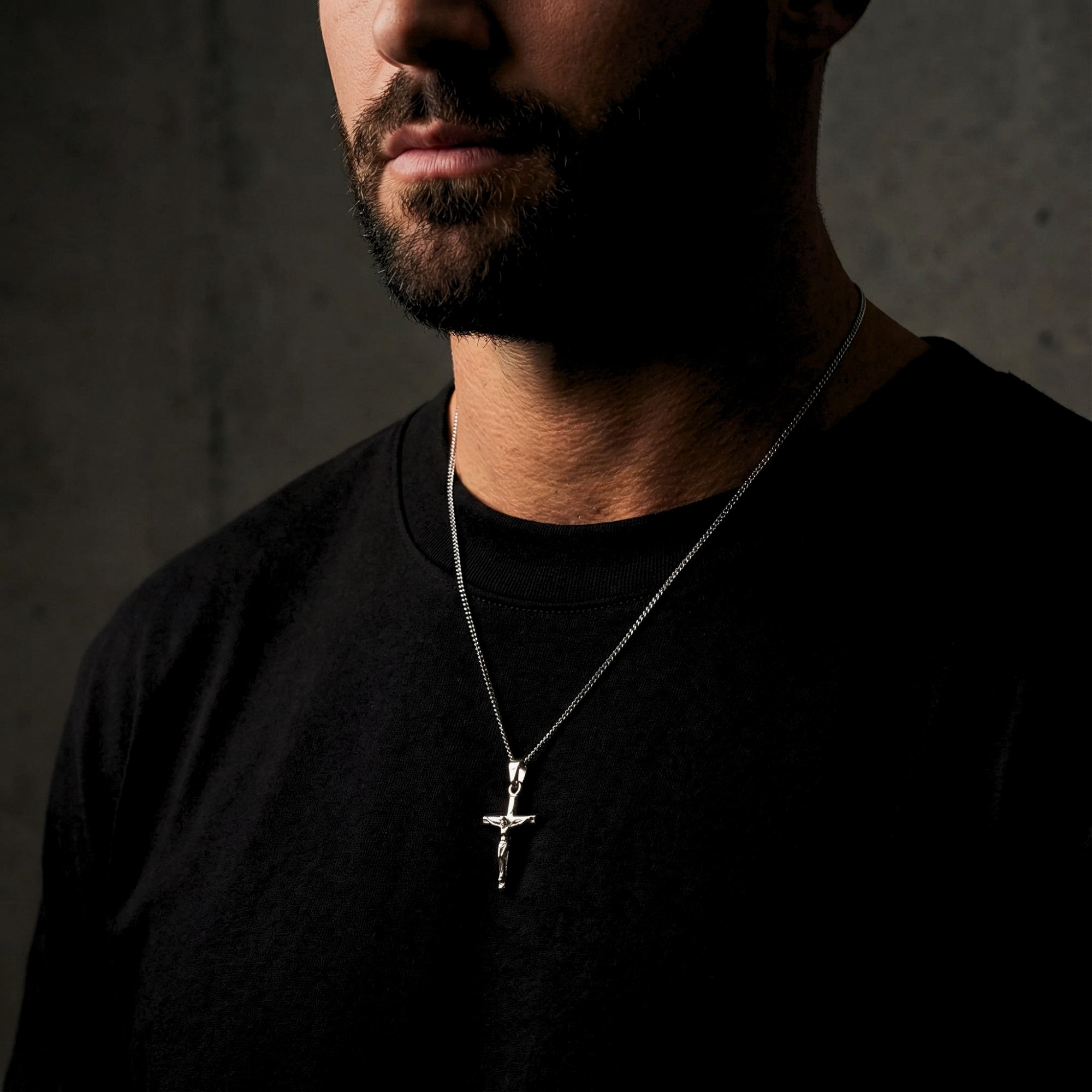 Men's silver stainless steel crucifix pendant necklace worn by a model over a black crew neck t-shirt.