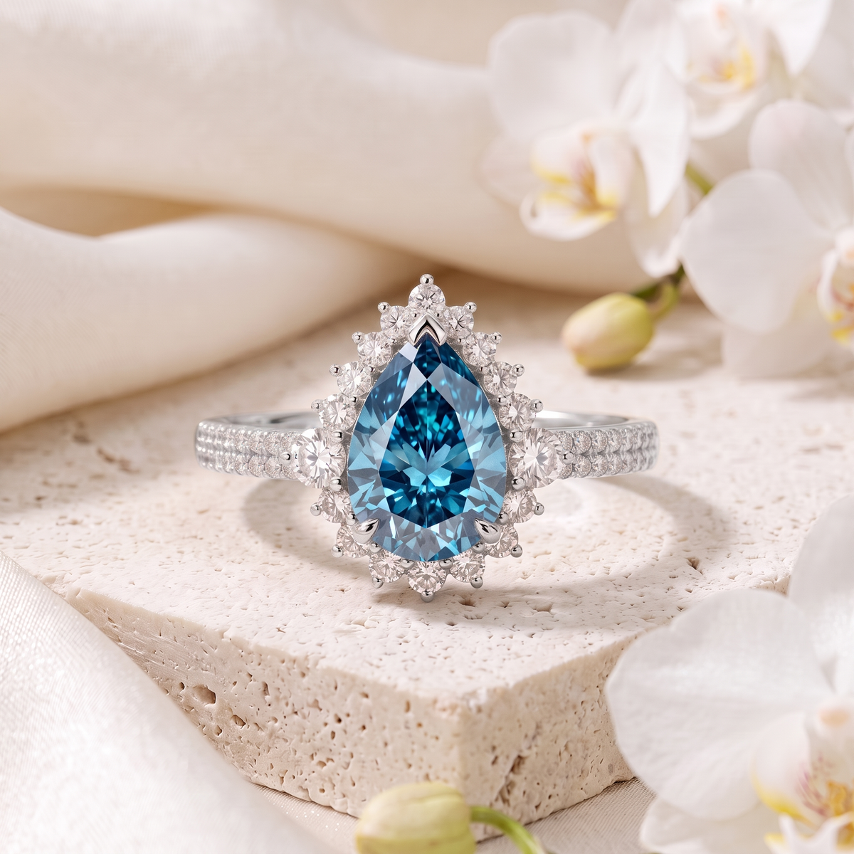 Pear cut blue moissanite halo engagement ring with a double pavé band in white gold resting on a stone display.