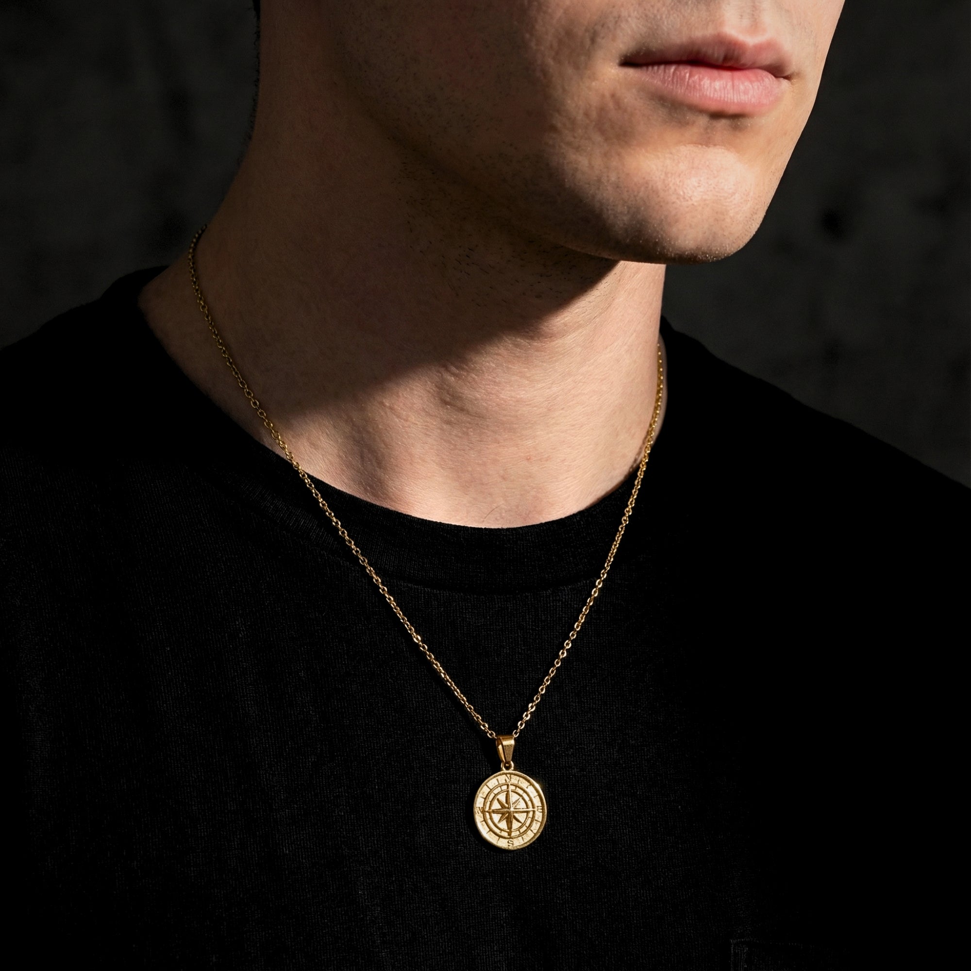 Gold stainless steel Voyager compass pendant necklace worn by a man against a dark background.