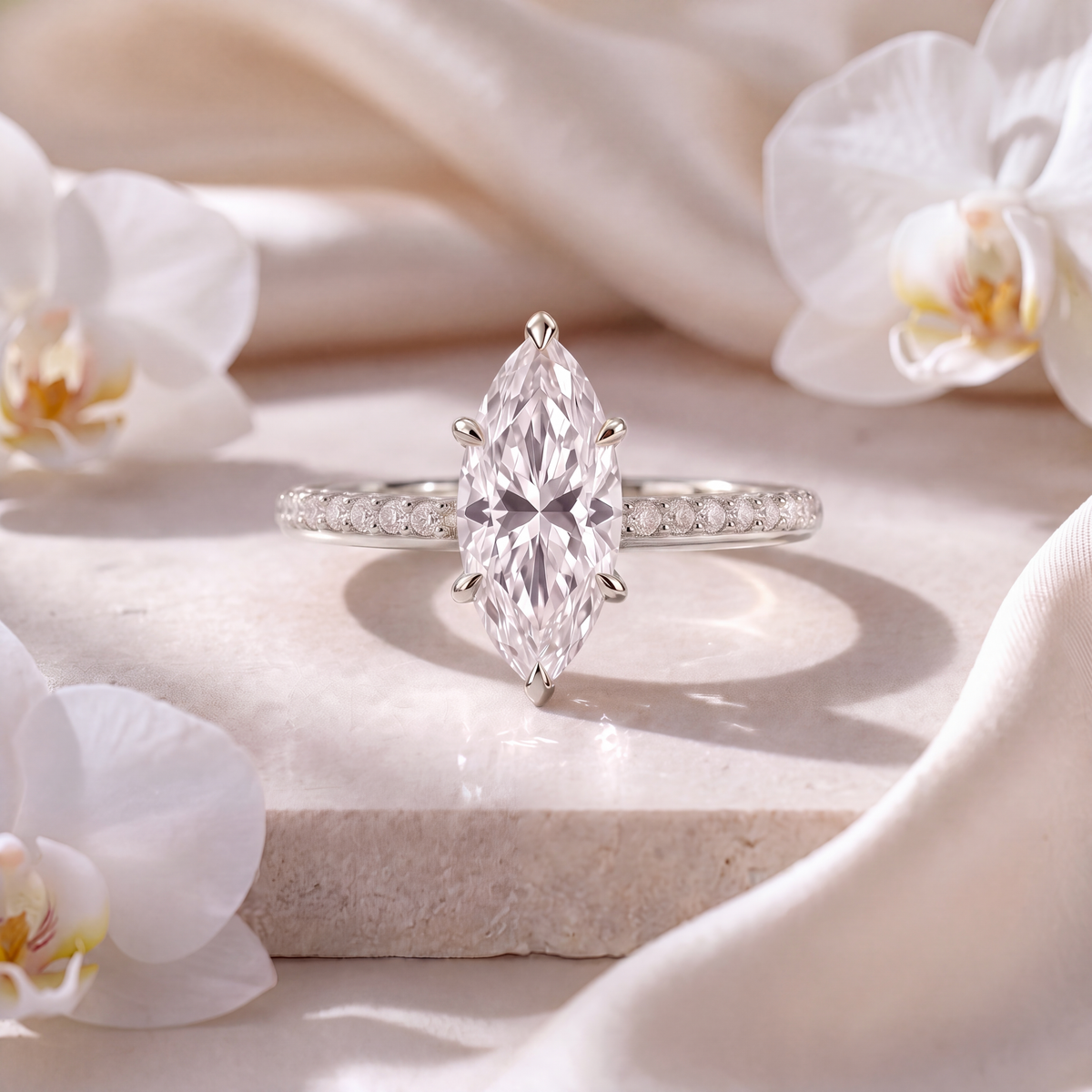 White gold marquise moissanite engagement ring with a pavé band displayed on a marble surface.