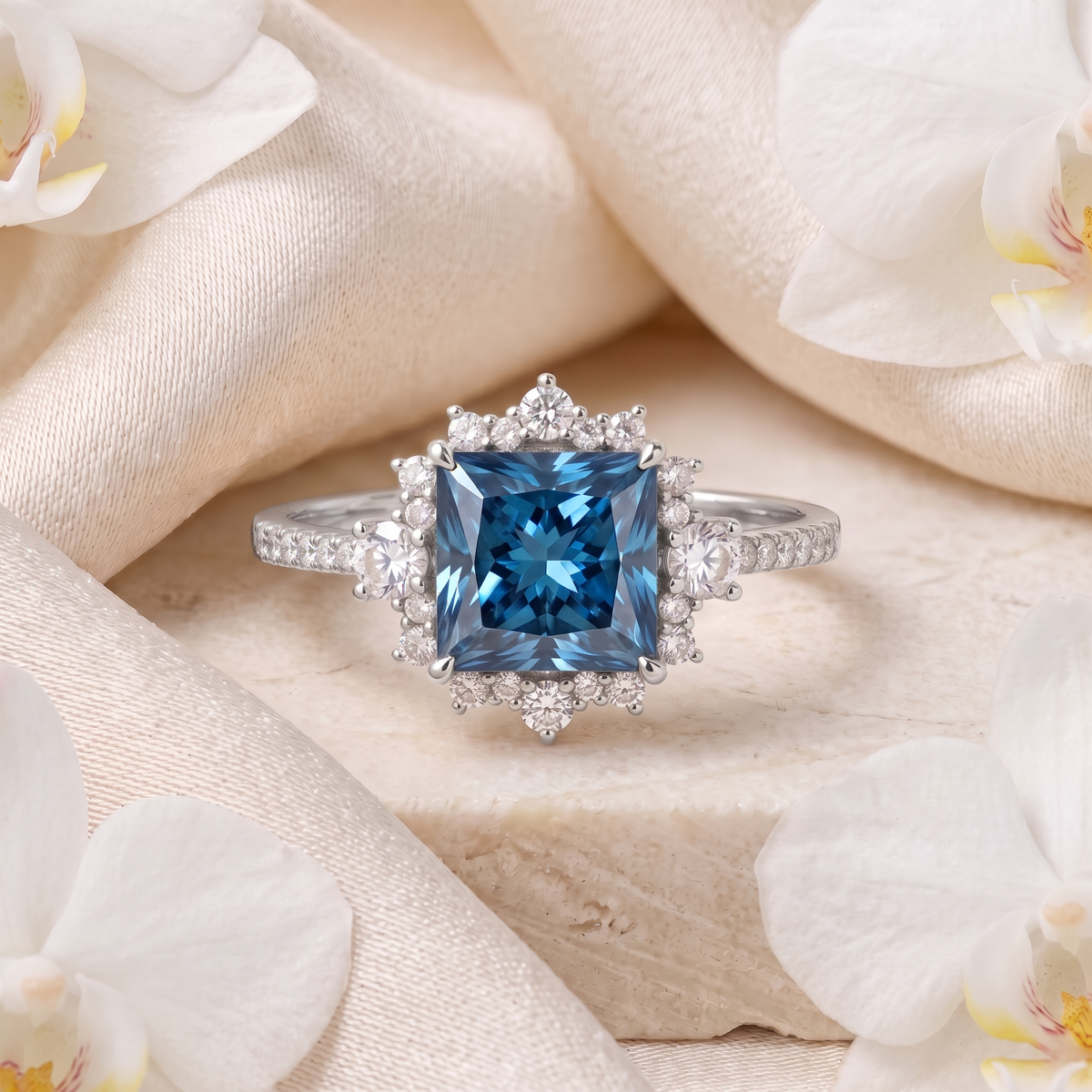 White gold princess cut blue gemstone ring with halo accents on a satin and marble background.