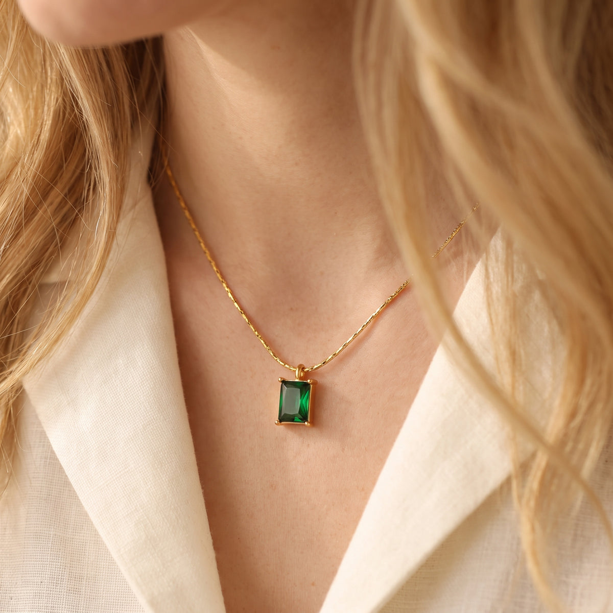 Front close-up of a gold-tone necklace with a rectangular emerald-green pendant worn with a white shirt.