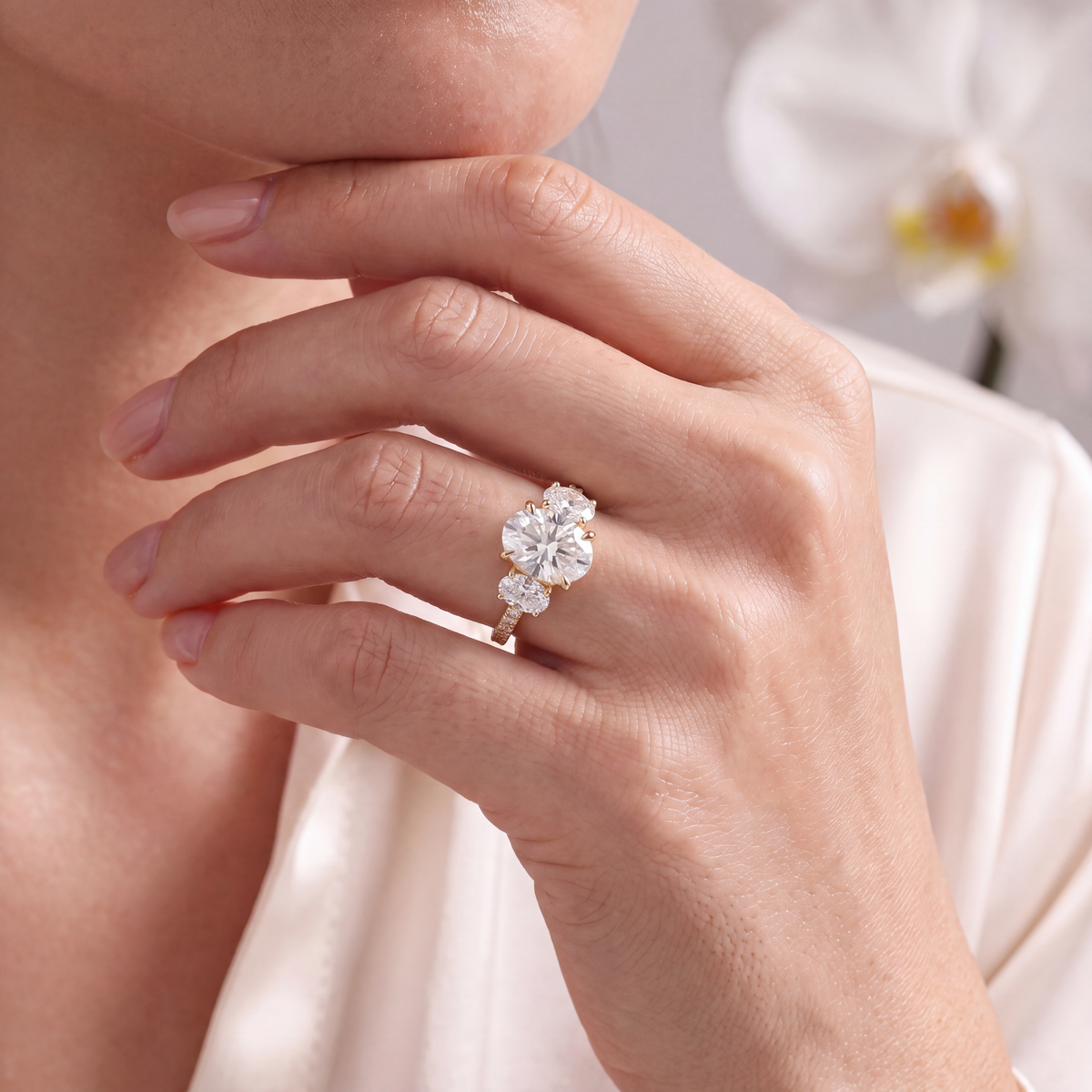 Close-up of a model holding her hand near her chin wearing the yellow gold three-stone oval moissanite ring.