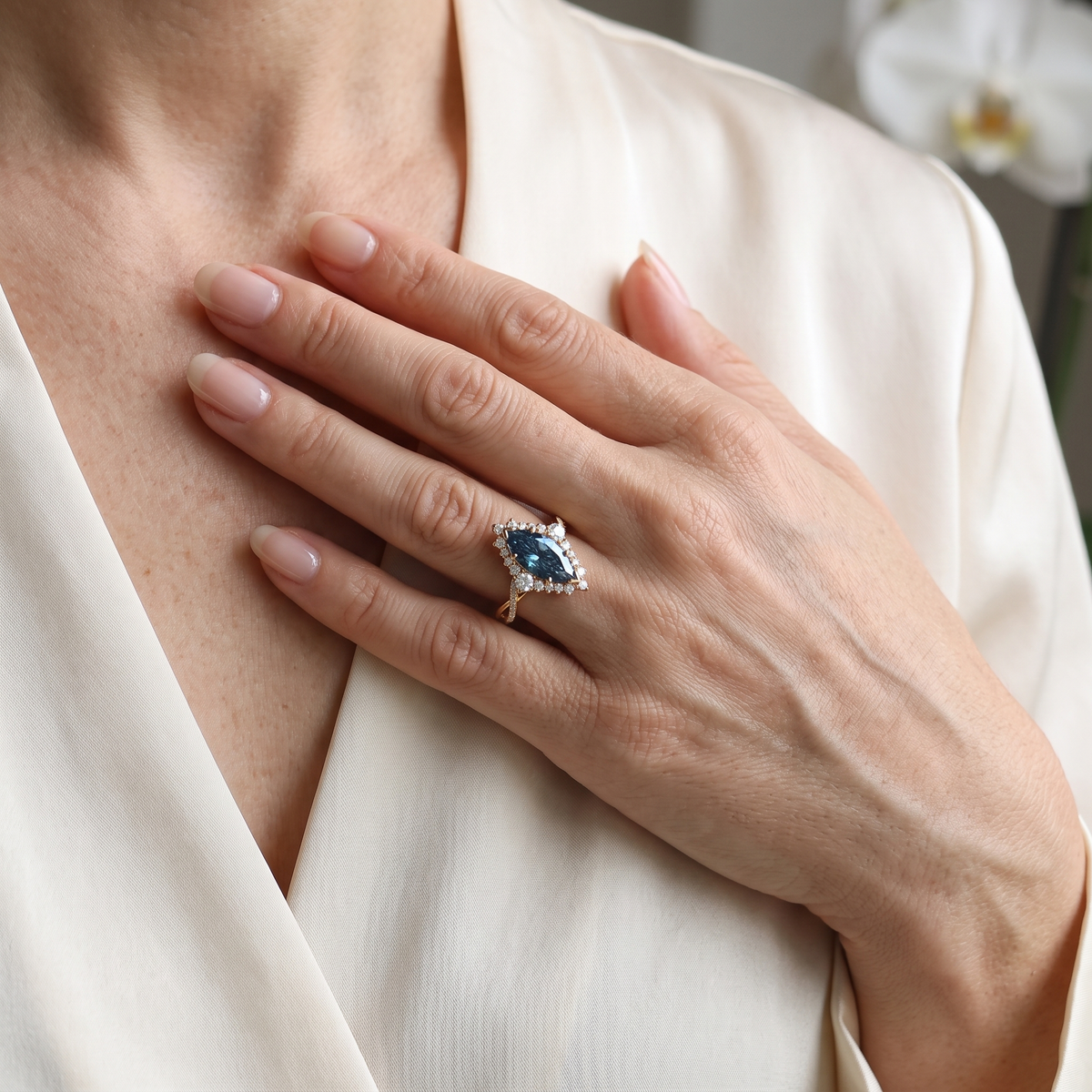 Lifestyle view of a model resting her hand on her chest while wearing a yellow gold blue marquise moissanite halo ring.