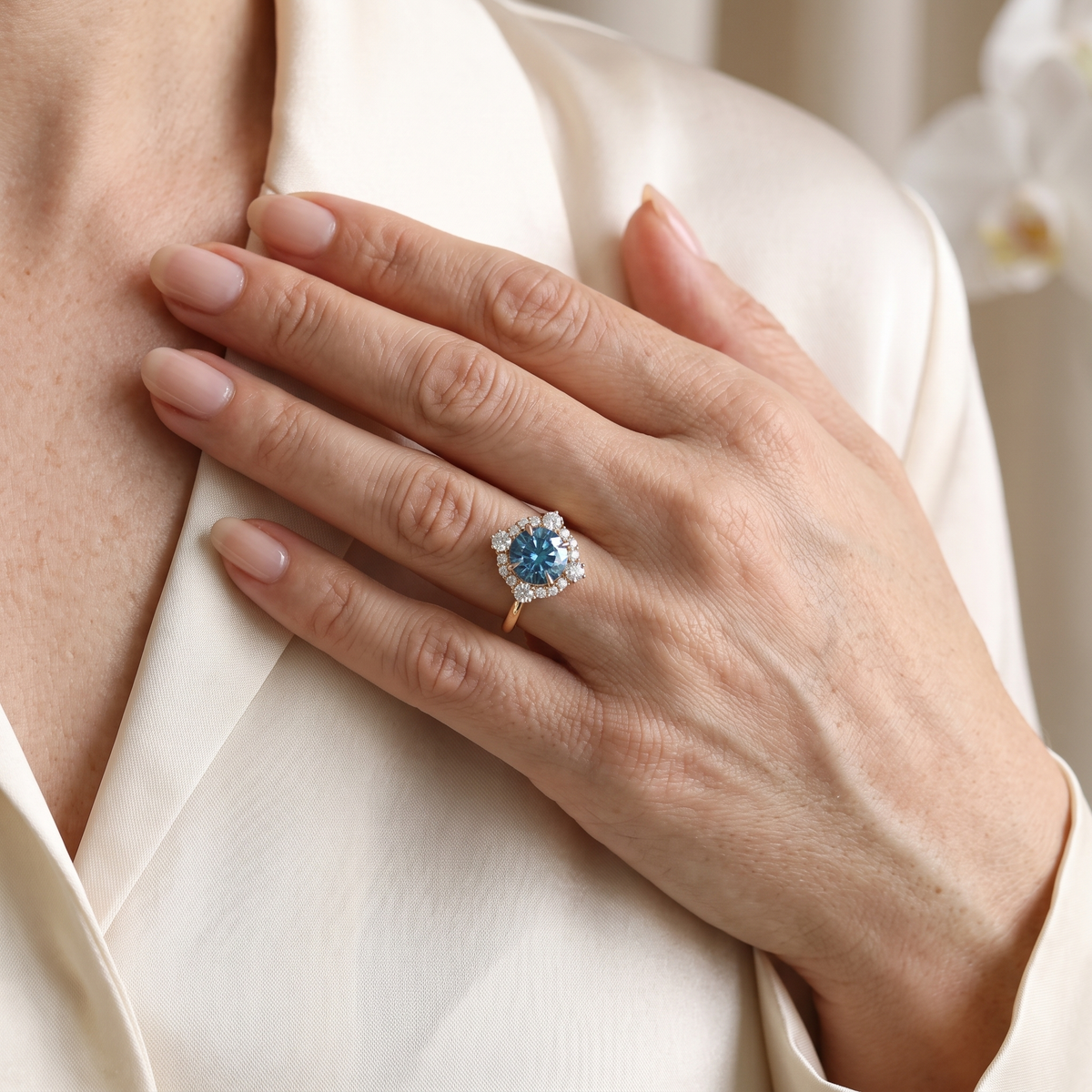 Close-up of a rose gold blue moissanite halo engagement ring worn on a hand against ivory silk.