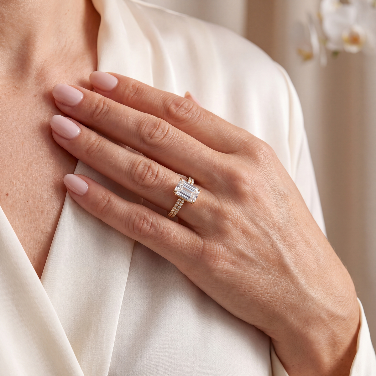Model placing hand on chest showing gold emerald-cut moissanite engagement ring with double-row pavé band.