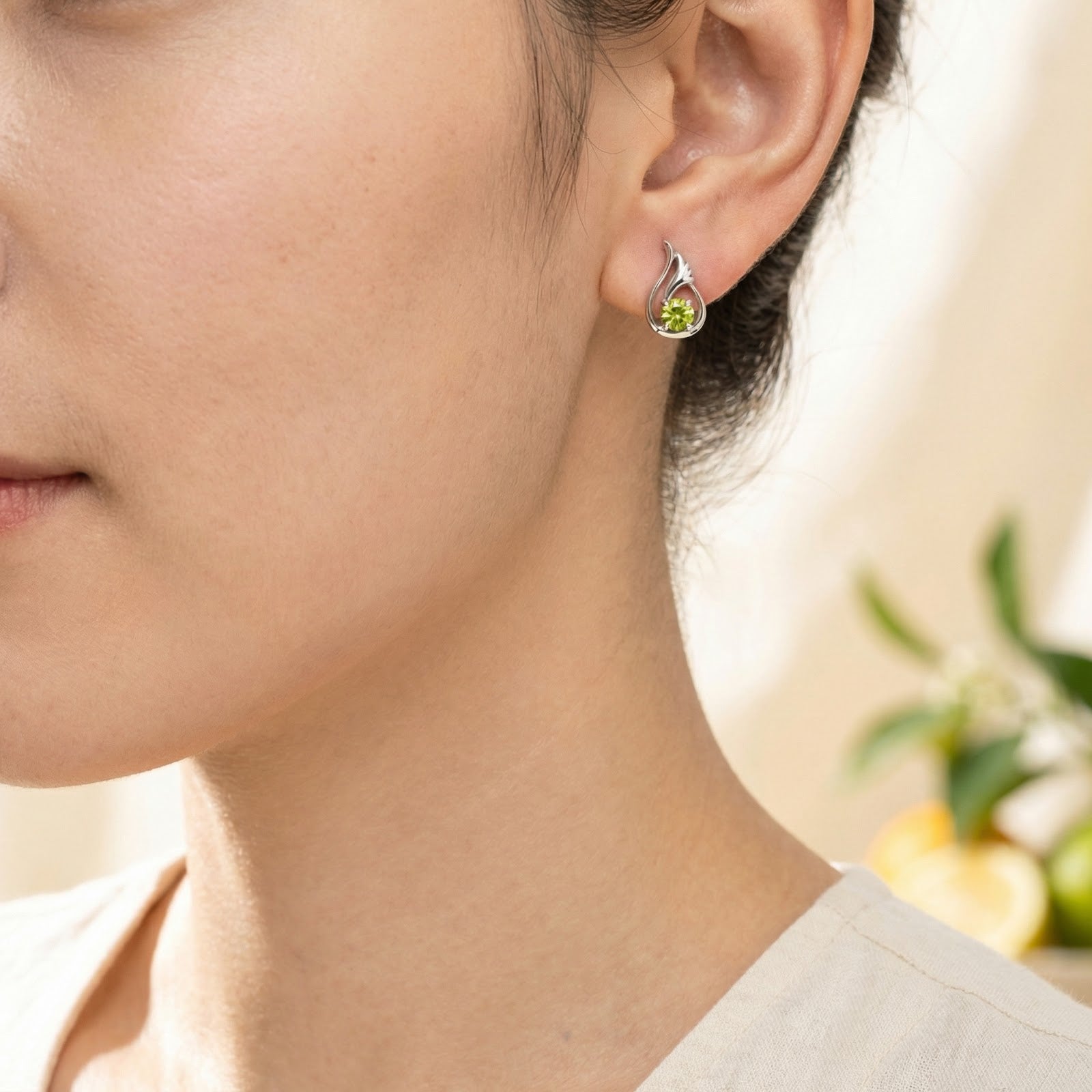 Close-up of a silver tulip bud earring with a round green simulated peridot stone worn on a model's ear.