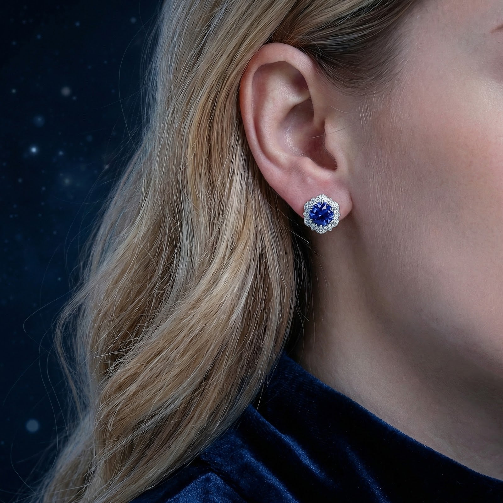 Close-up of a round blue simulated sapphire halo stud earring in S925 sterling silver worn on a woman's ear.