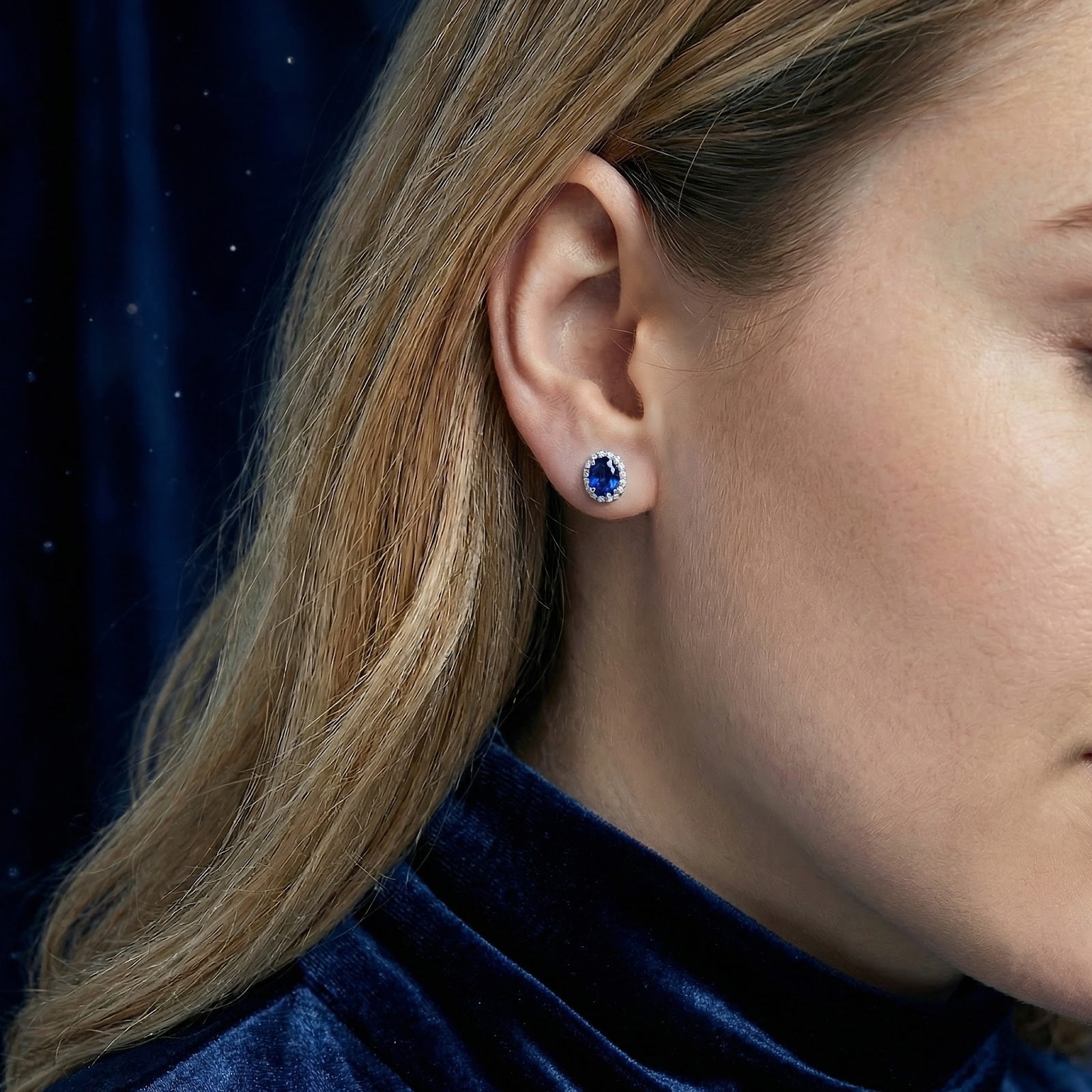 Side profile of a woman wearing an oval simulated sapphire halo stud earring in sterling silver.