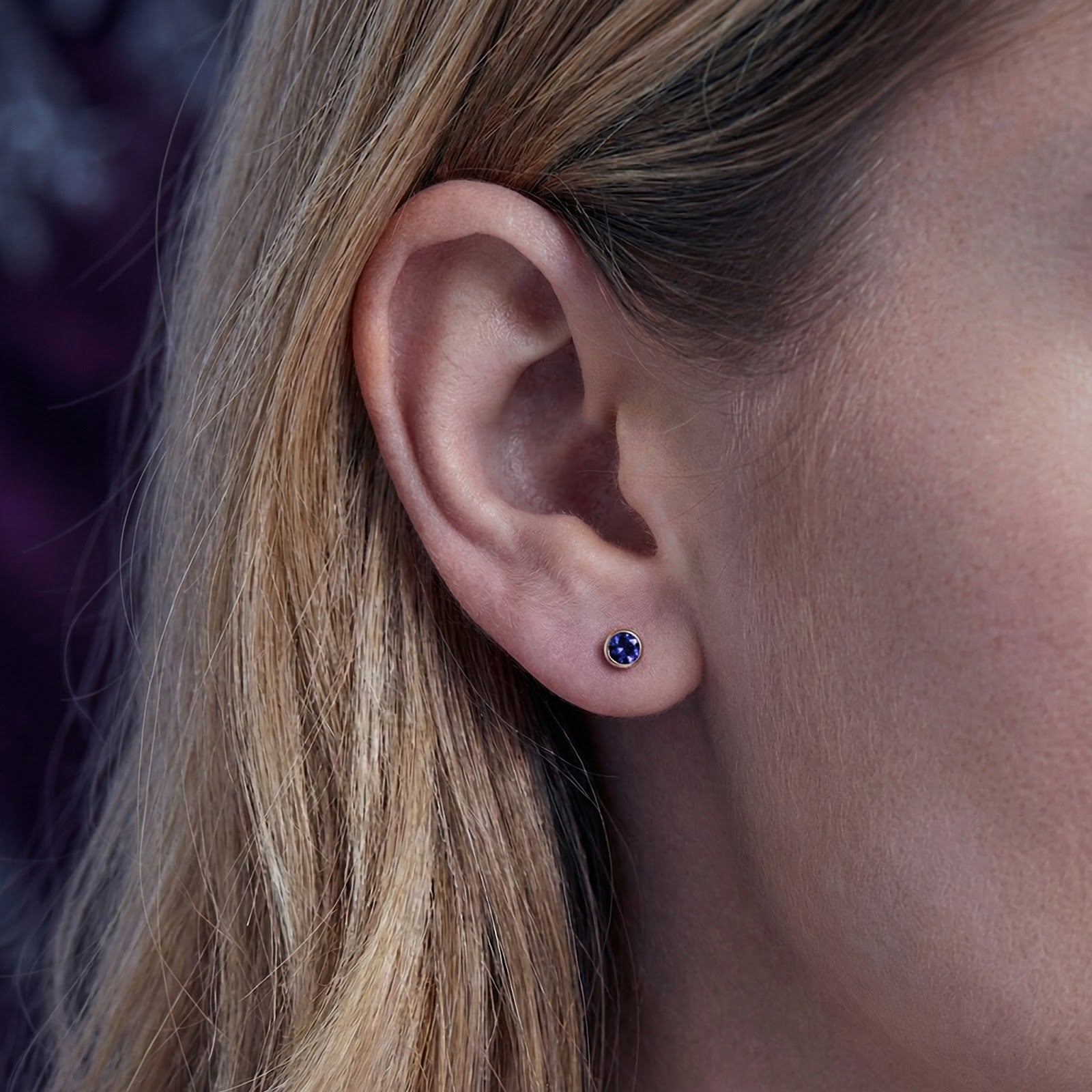 Close-up of a woman wearing a gold vermeil bezel stud earring with a round simulated tanzanite stone.