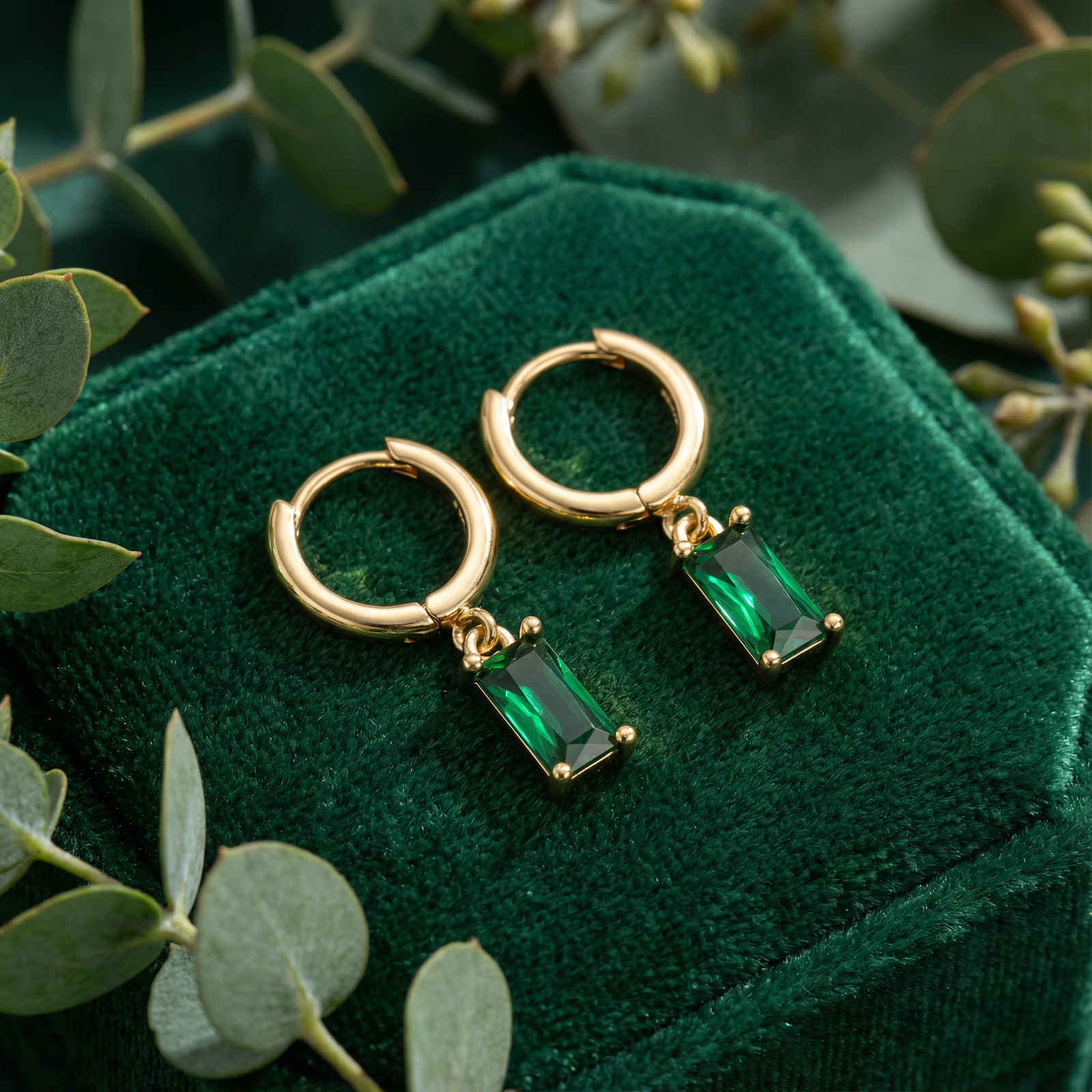 Gold vermeil hoop earrings with emerald-cut simulated emerald drops on a green velvet box with eucalyptus.