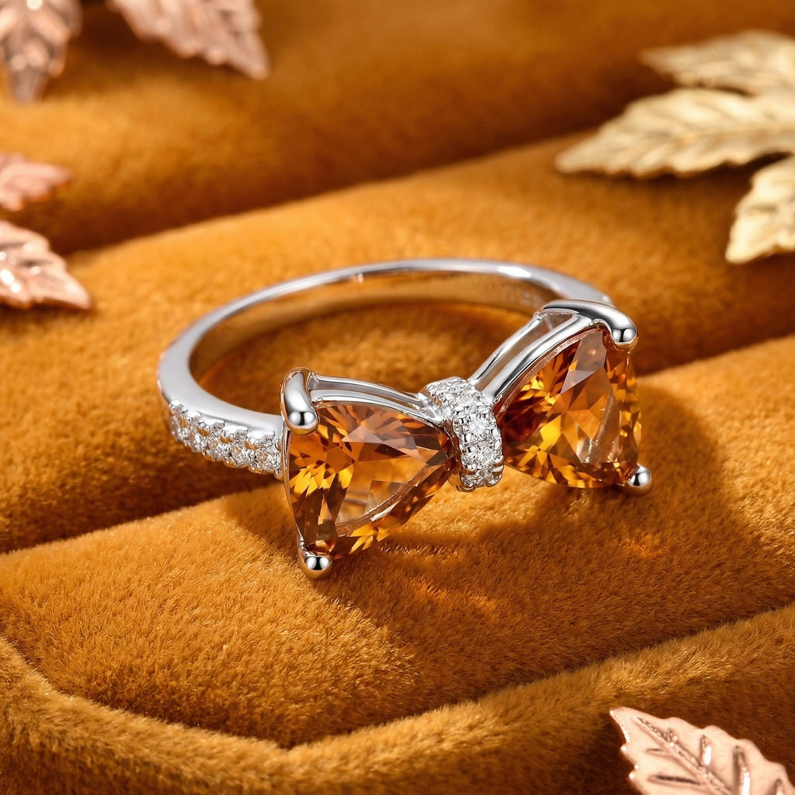 Front view of a sterling silver bow ring with trillion-cut honey topaz stones in a velvet box with fall leaves.