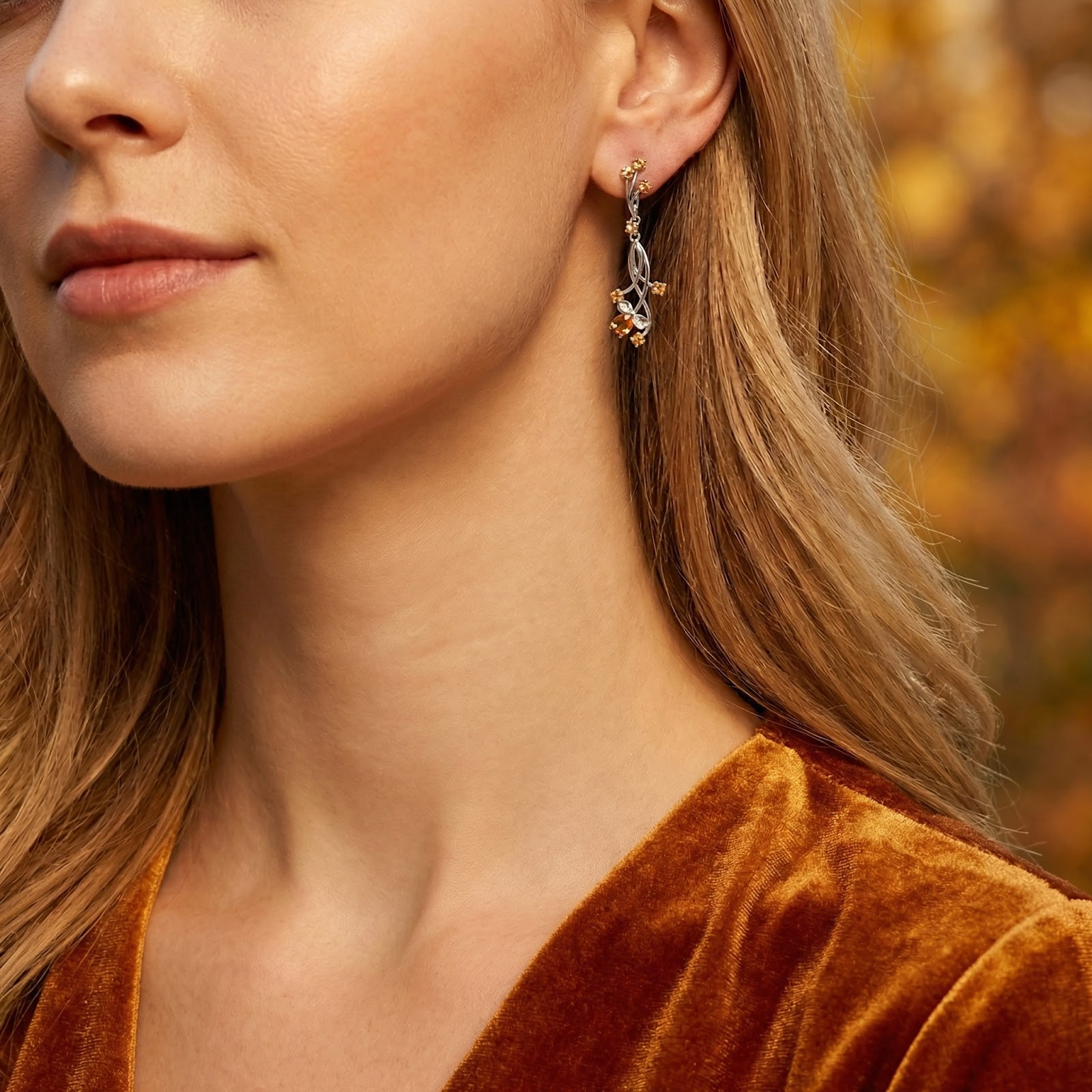 Woman wearing a sterling silver vine drop earring with simulated topaz stones against a blurred fall background.