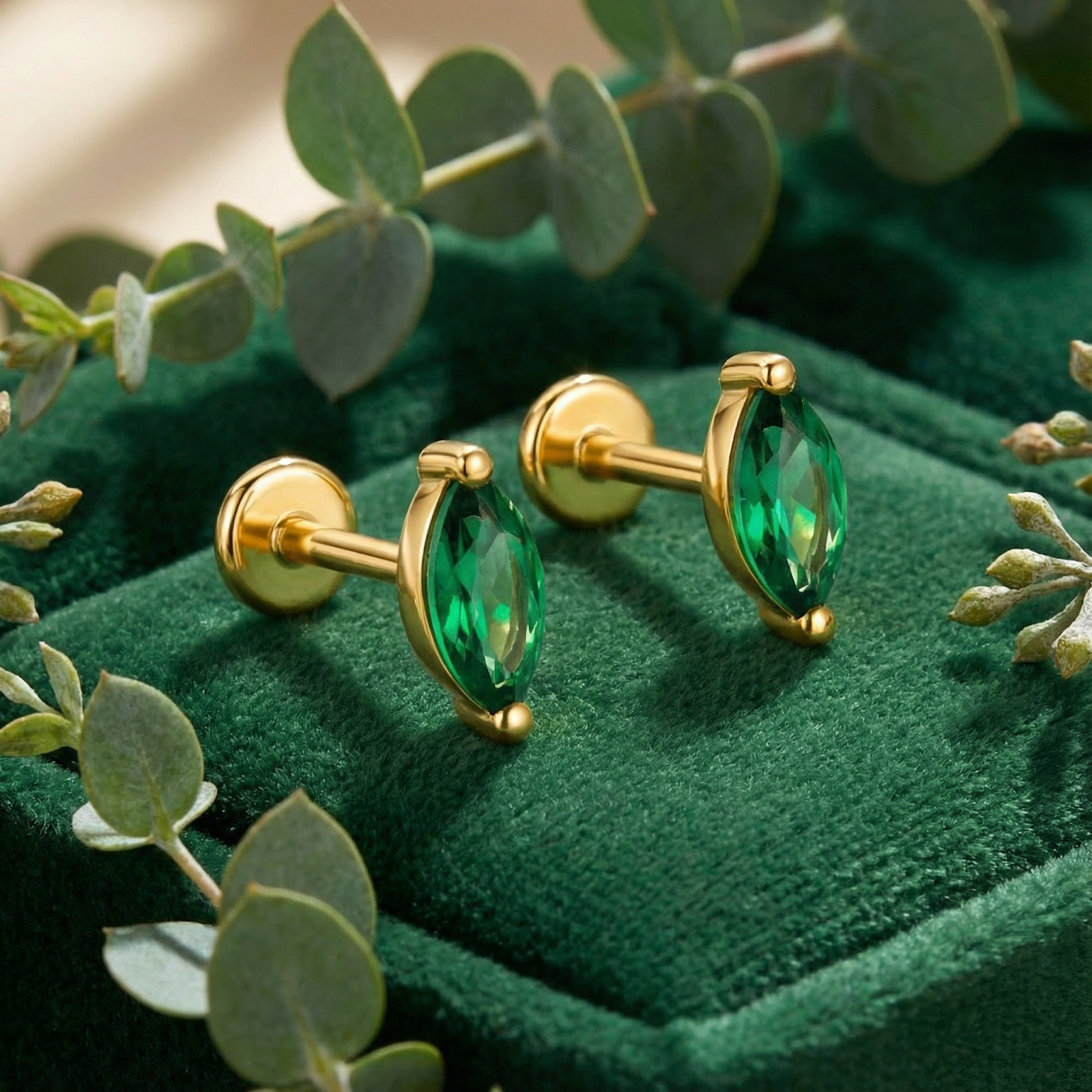 Marquise-cut simulated emerald stud earrings in 18K gold vermeil on green velvet with eucalyptus leaves.
