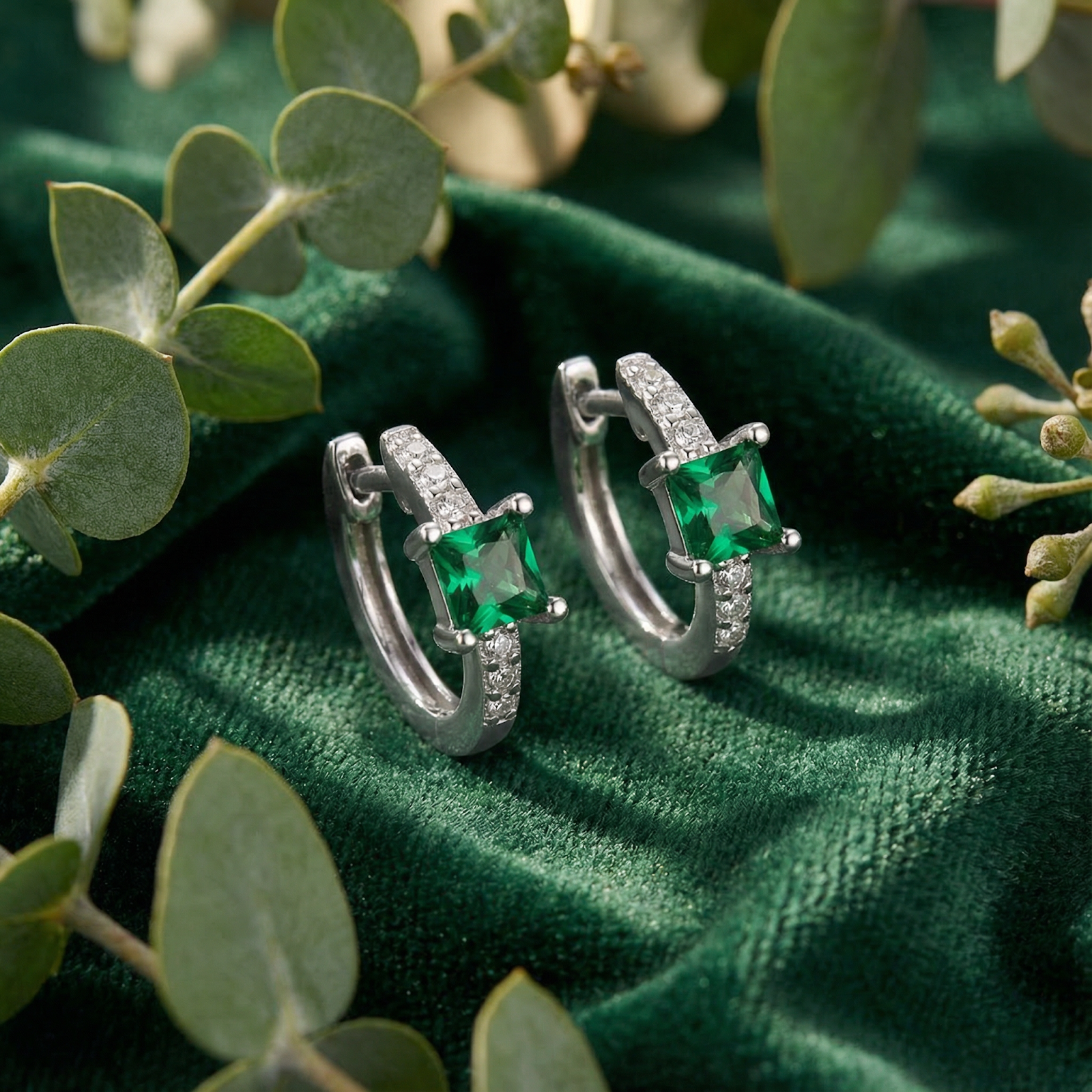Sterling silver huggie earrings with square-cut simulated emeralds on green velvet with eucalyptus leaves.