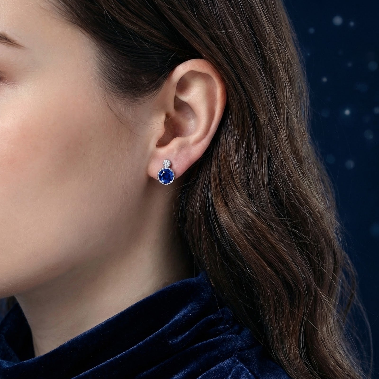 Woman with dark hair wearing a round blue simulated sapphire and sterling silver halo stud earring.