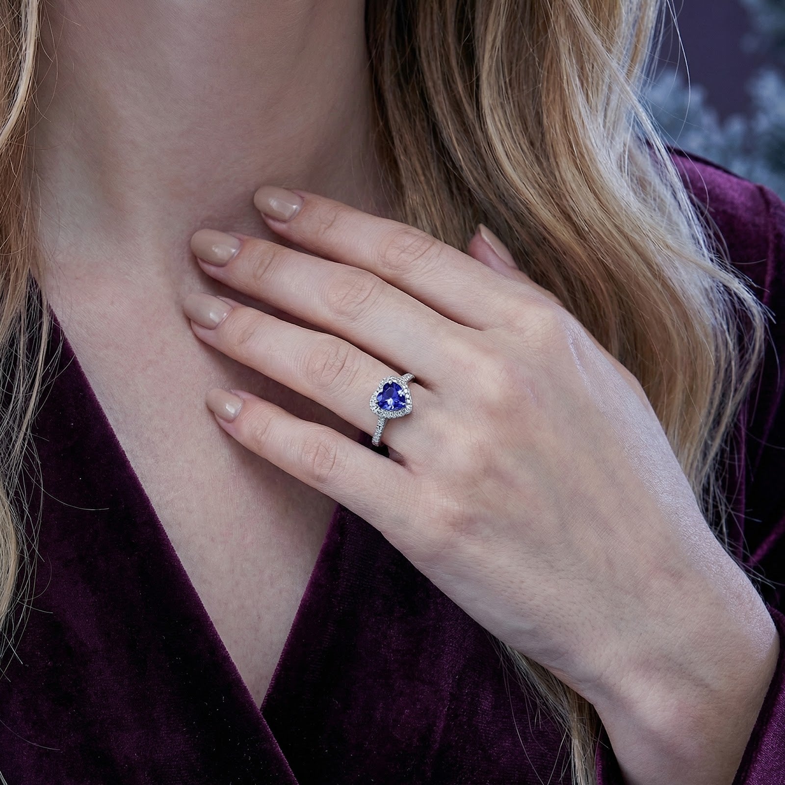 Heart-cut simulated tanzanite halo ring in sterling silver worn on a woman's hand against a purple dress.