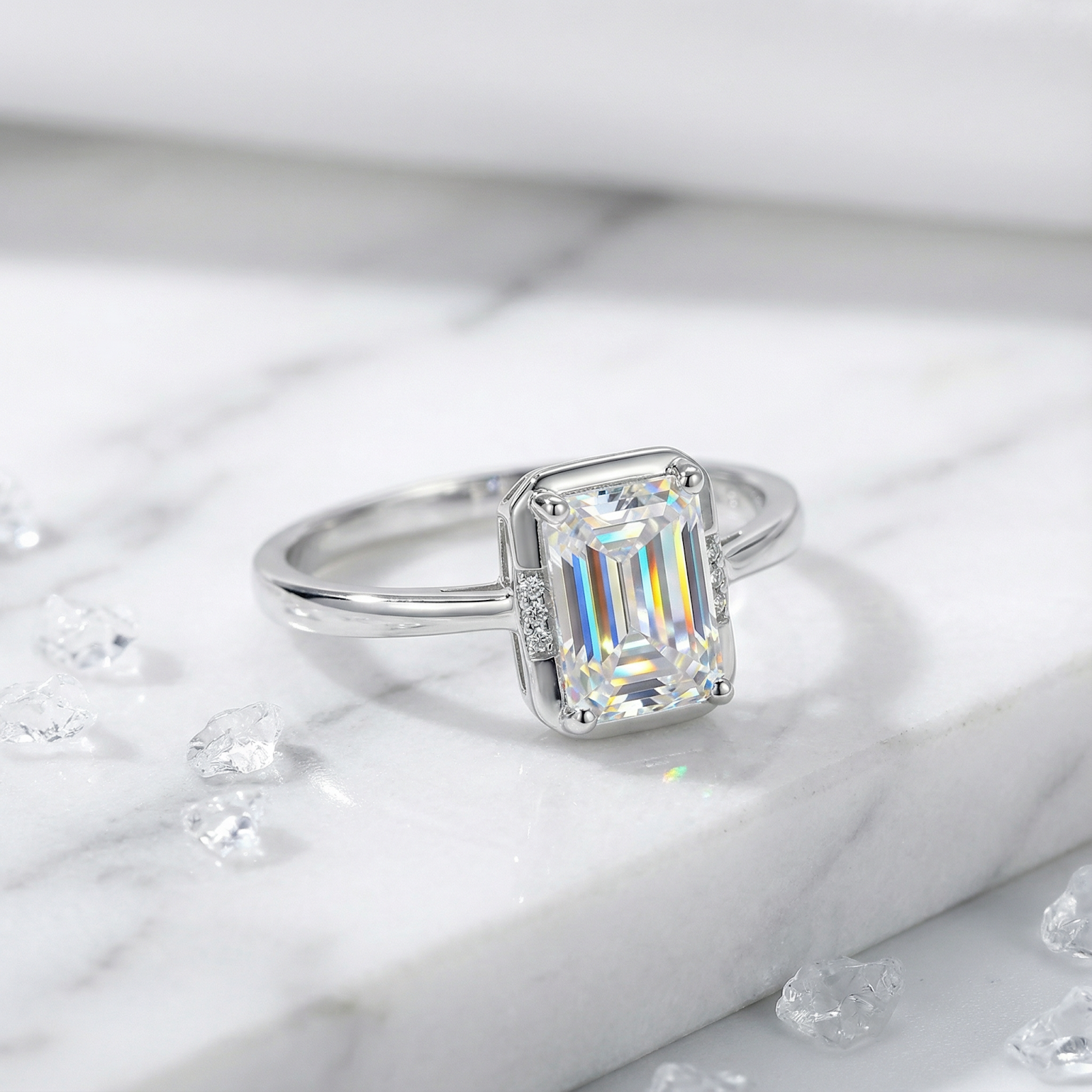 Emerald-cut moissanite ring in sterling silver displayed on a marble surface with loose crystals.