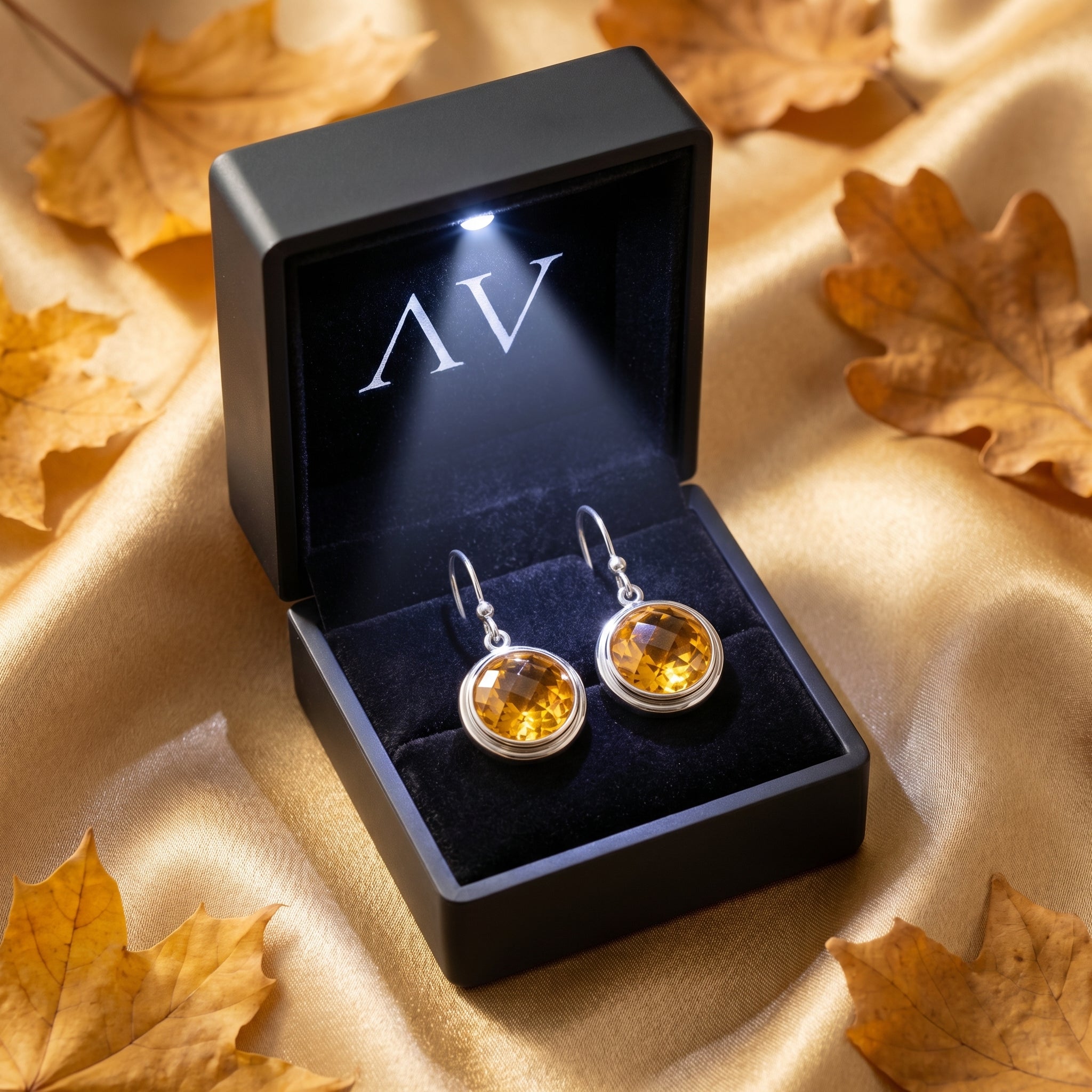 Round-cut citrine drop earrings presented in a lighted black jewelry box on a golden background.