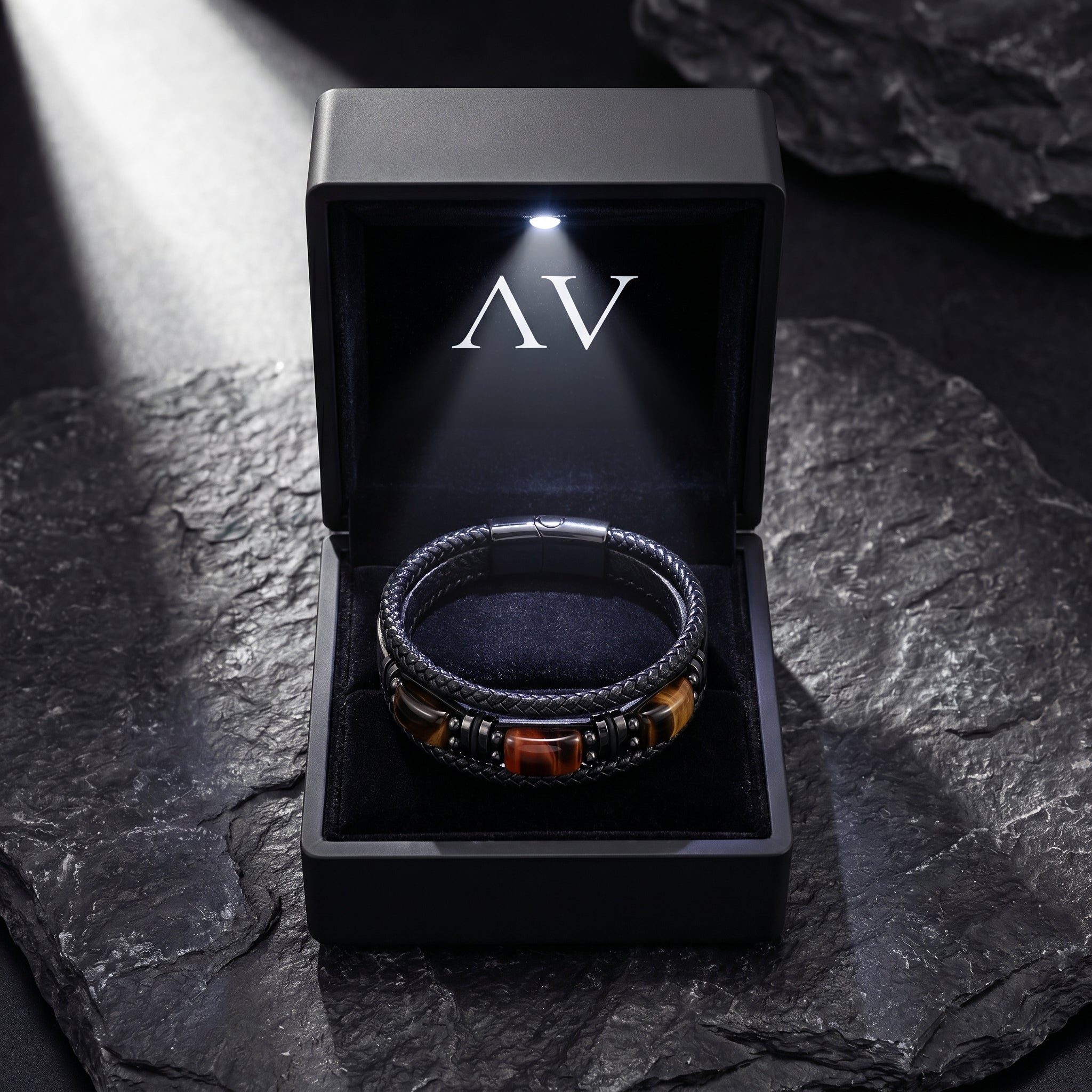 Men's black braided leather bracelet with tiger's eye stones in a black LED-lit presentation box on a dark rock.
