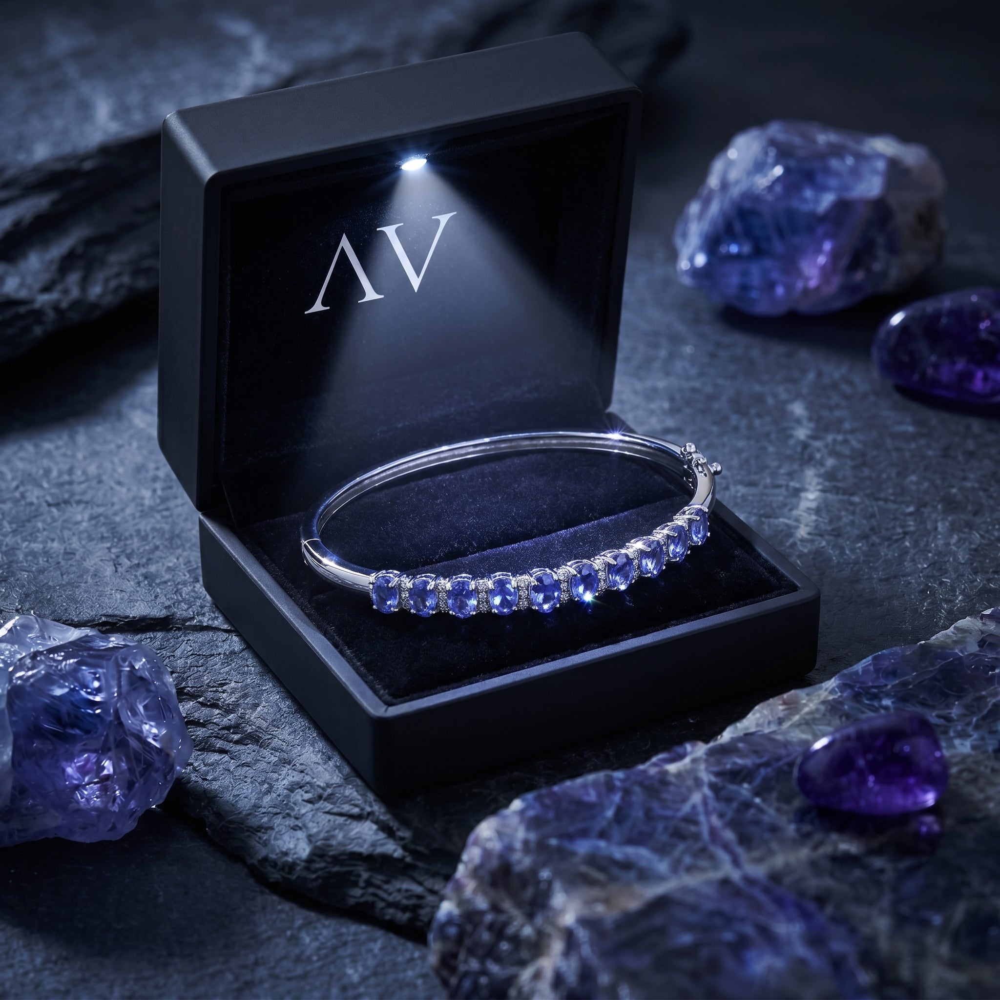 Silver oval tanzanite bracelet inside a premium black LED-lighted presentation box on a dark stone background.