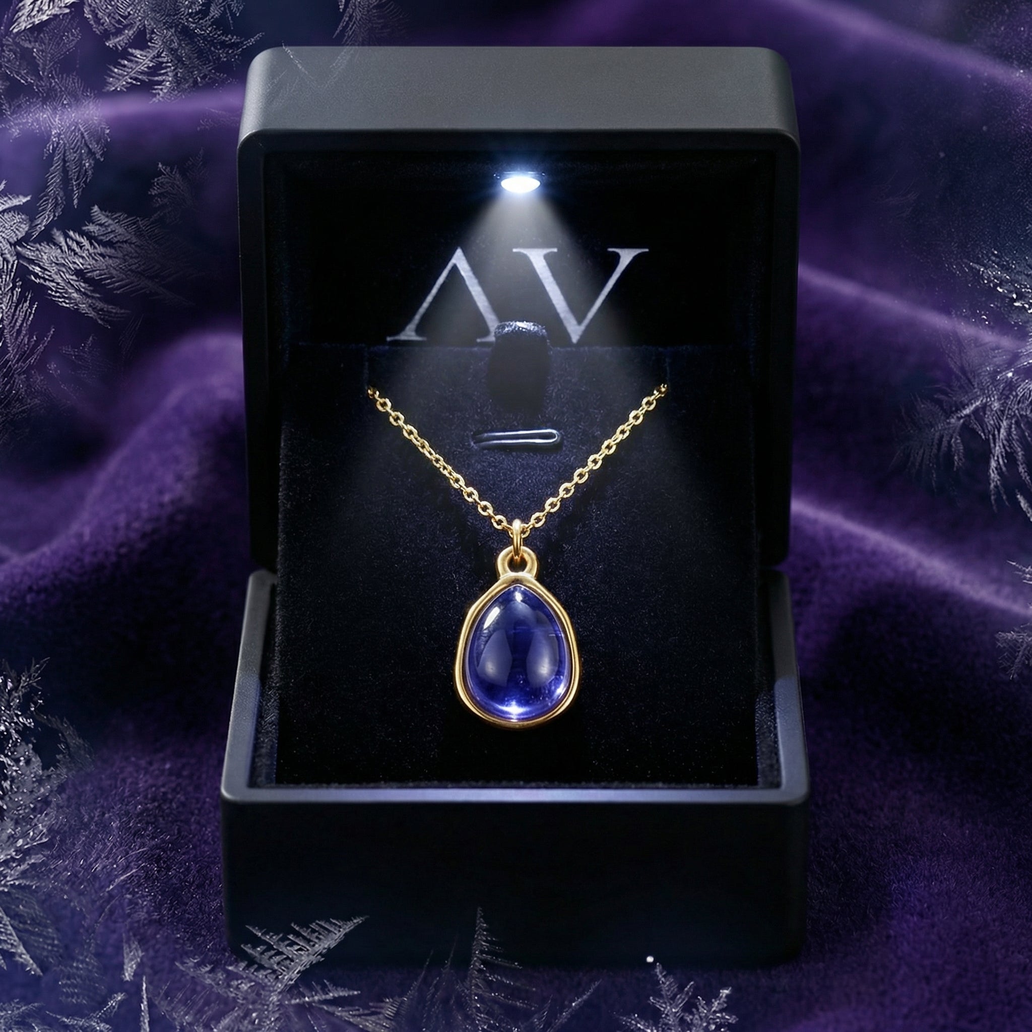 Gold teardrop simulated tanzanite necklace in a lighted black jewelry box on a purple background.