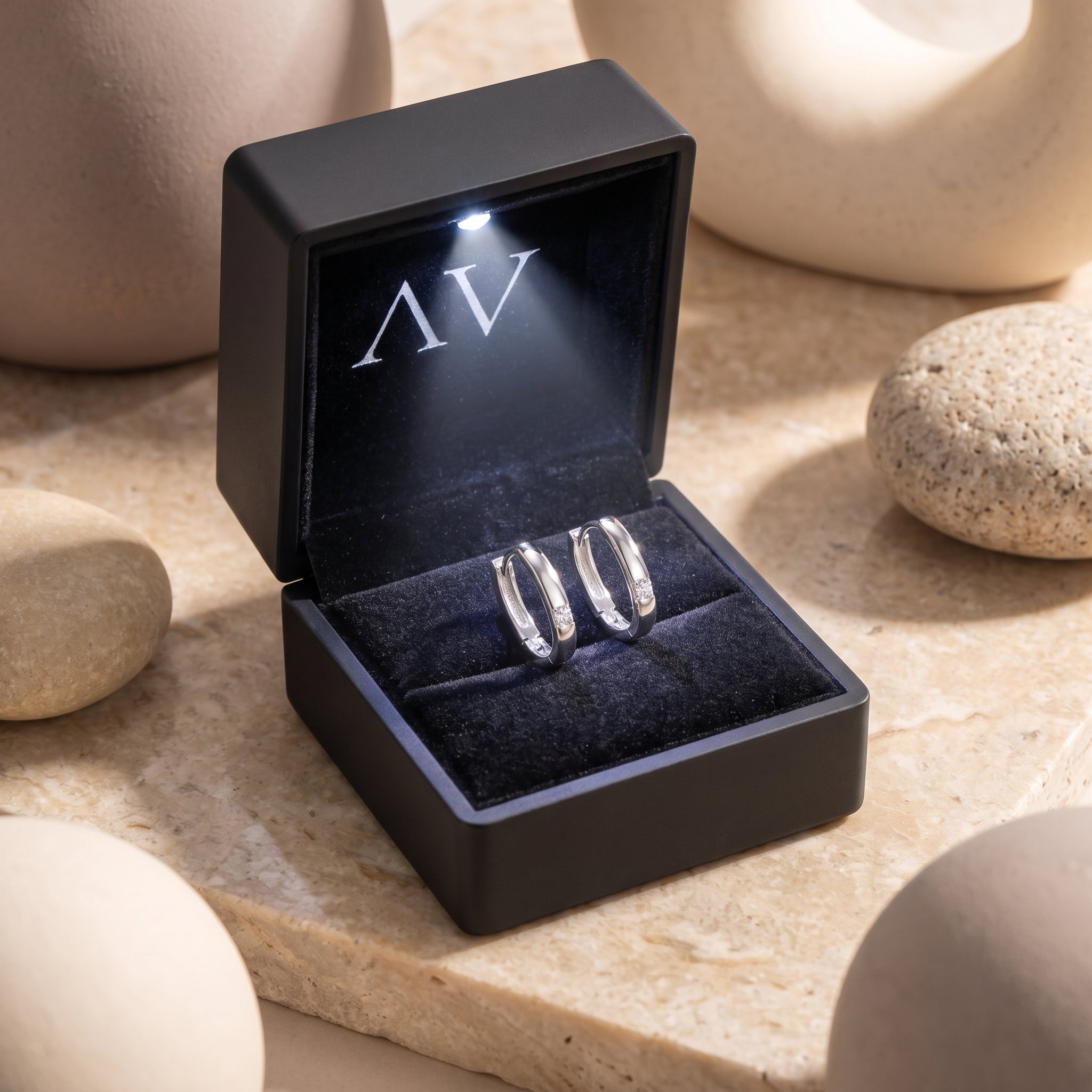 White gold round moissanite huggie hoop earrings presented in a black LED-lit jewelry box on a stone surface.