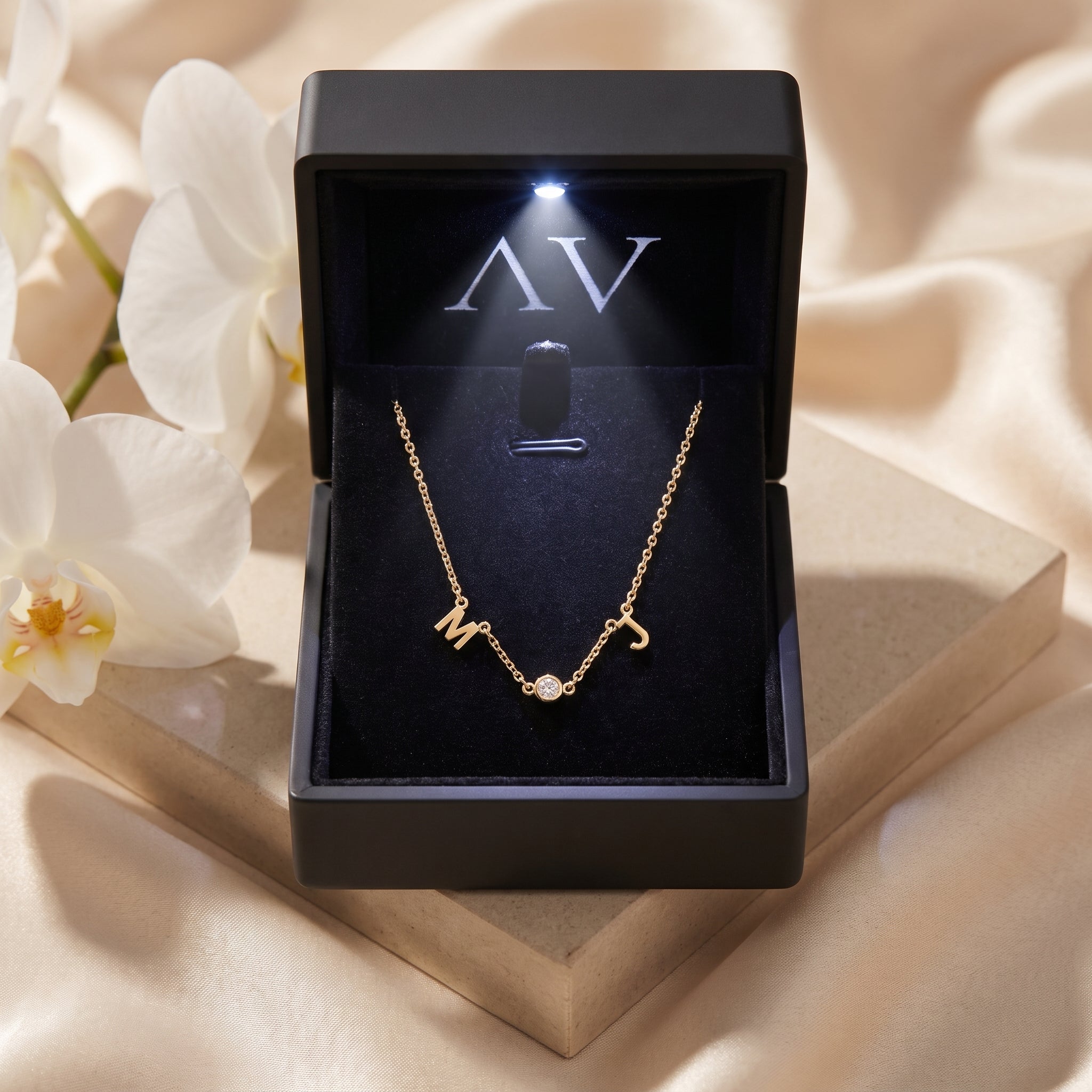 Gold initial necklace with M and J charms inside a black LED-lit jewelry presentation box.