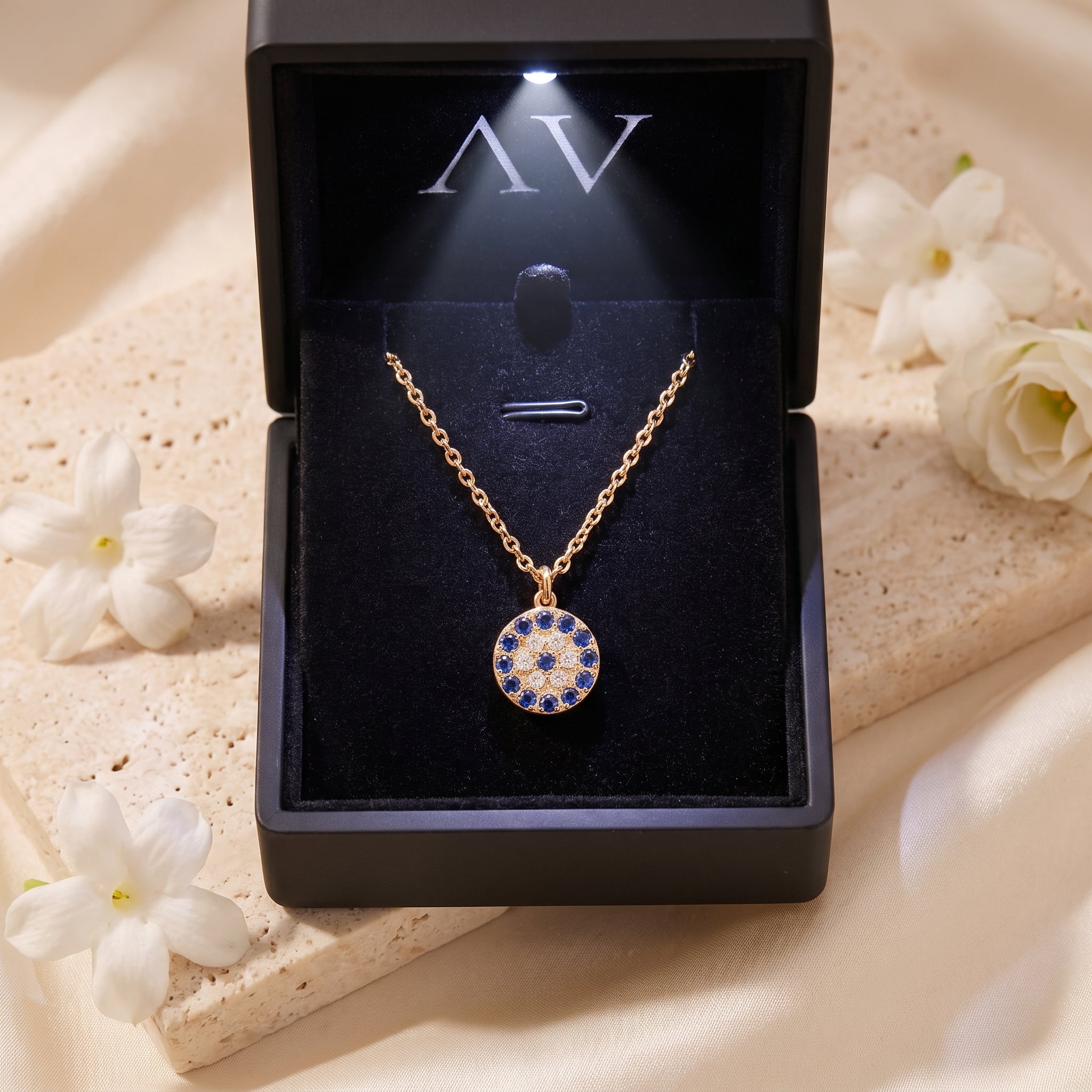 Gold PVD Stellis pendant necklace inside an illuminated black jewelry box with white flowers.