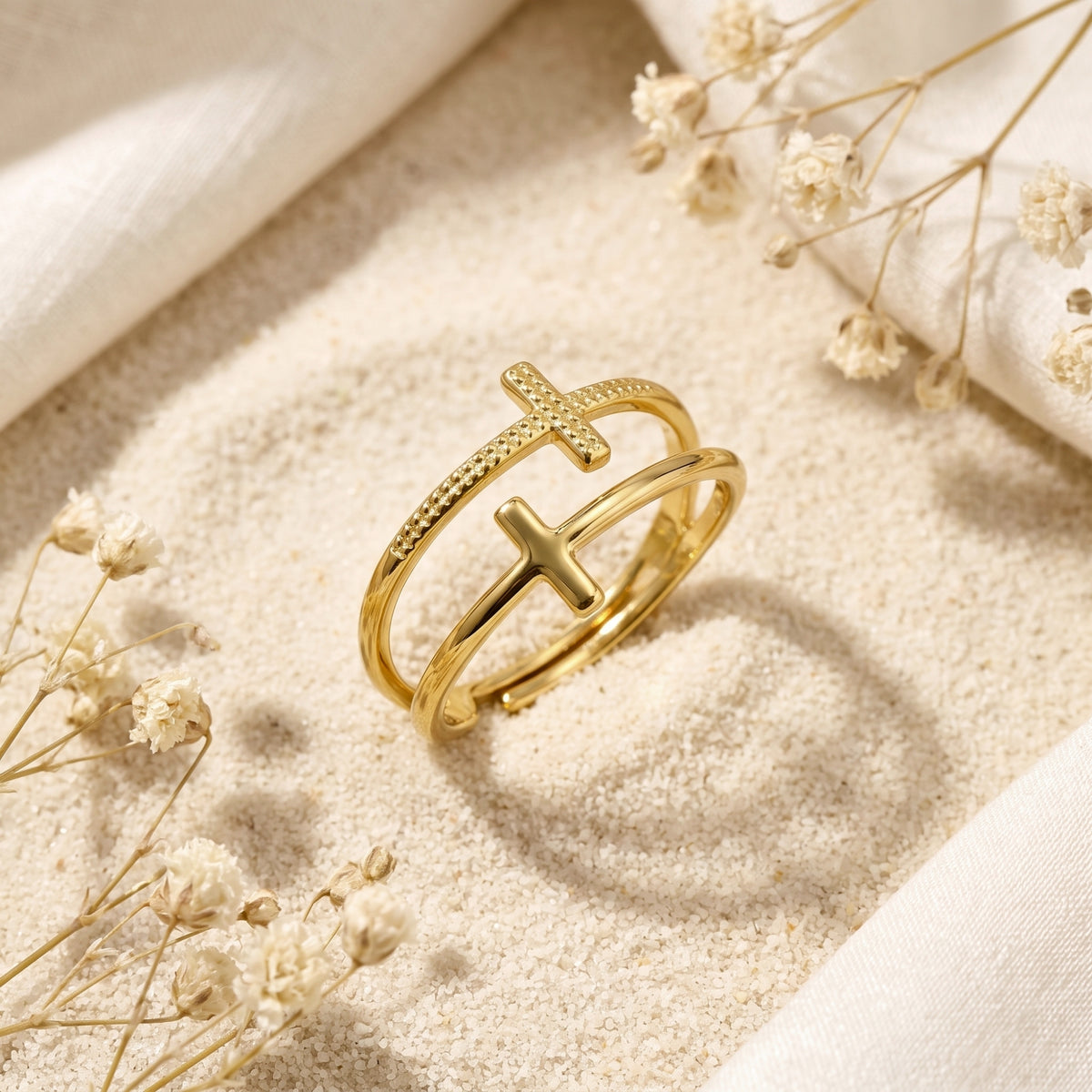 Gold-tone double cross ring displayed at an angle on fine sand with white dried flowers.