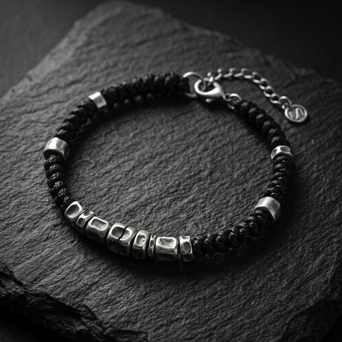 Black braided rope bracelet with sculpted silver beads, hook clasp, and extension chain on dark textured slate.