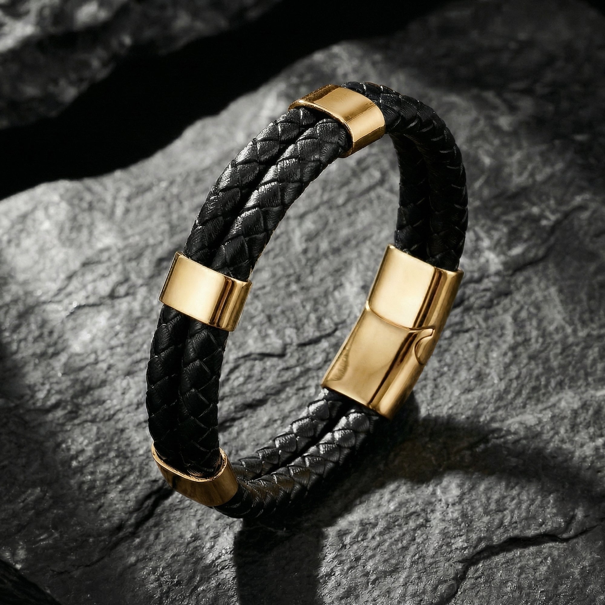 Men's braided black leather bracelet with gold stainless steel accents resting on a dark textured rock.