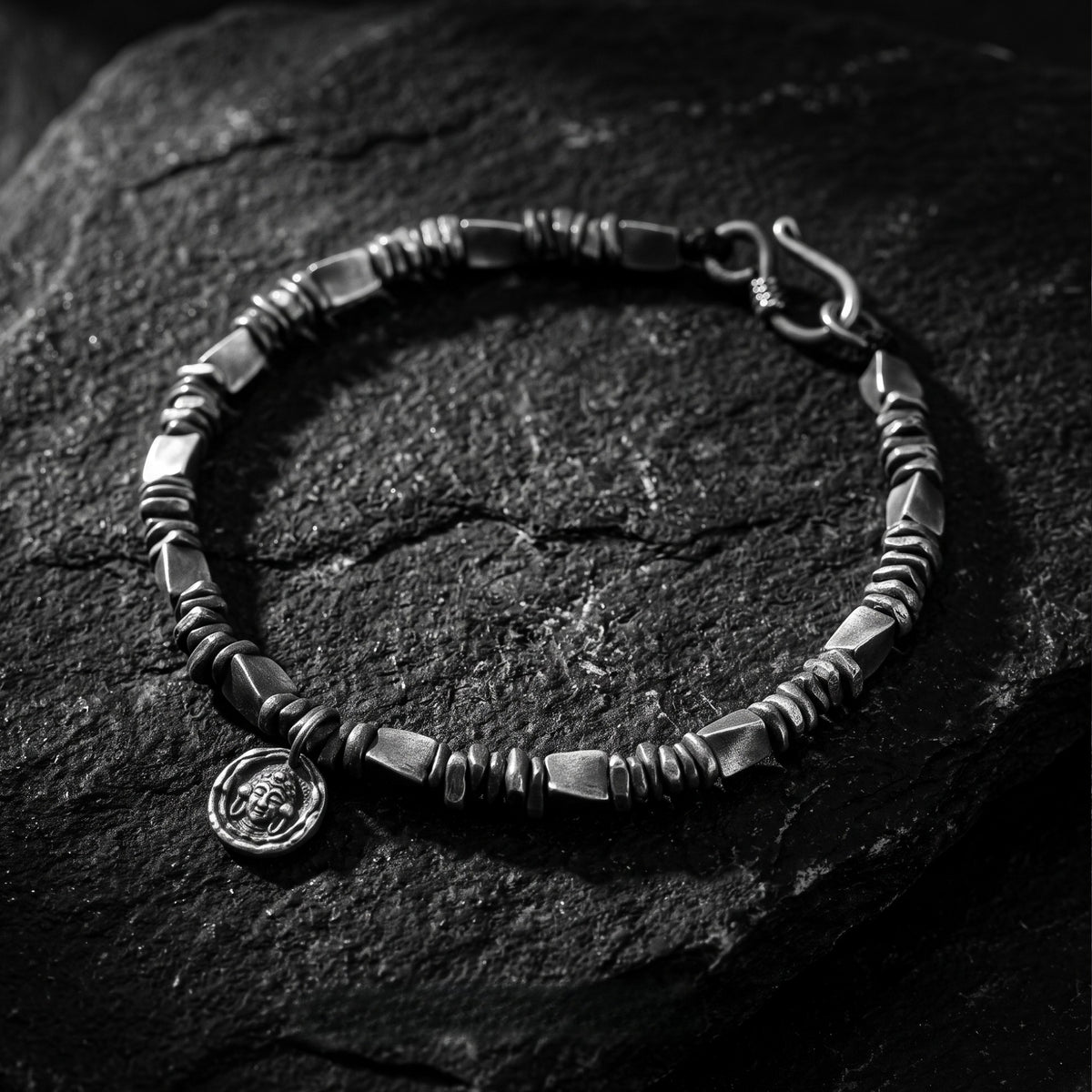 Angled close-up of a silver textured link bracelet with carved medallion resting on dark slate.