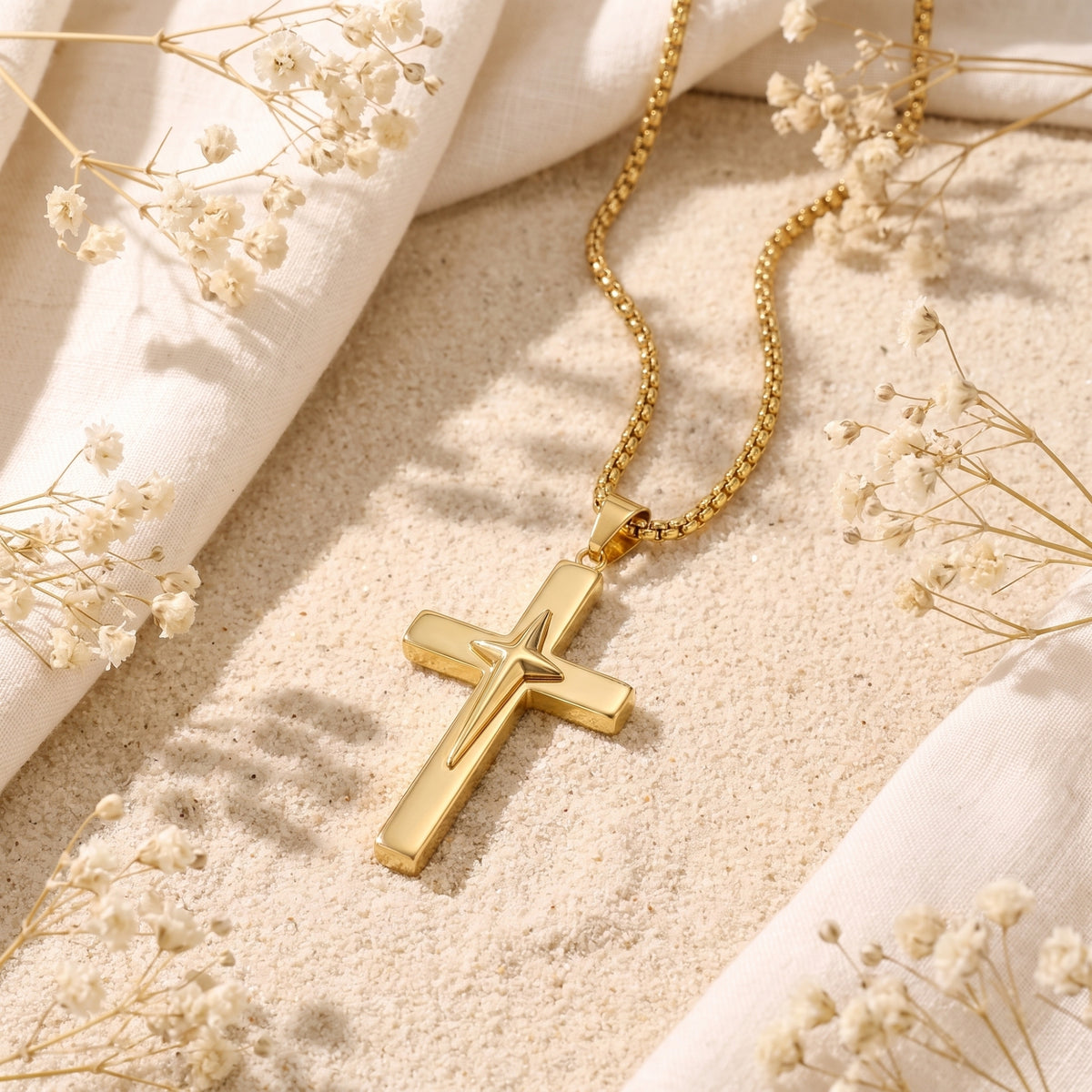 Gold-tone star cross necklace resting on sand beside white fabric and dried white flowers.