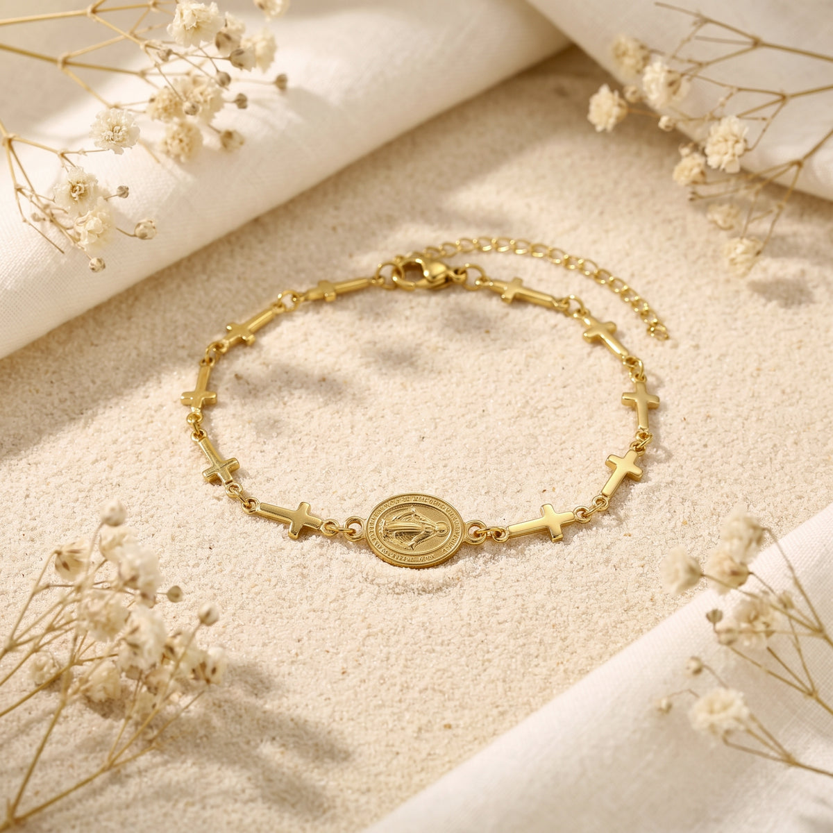 Overhead flat lay of a gold-tone cross link bracelet with an oval medallion on sand, surrounded by dried flowers.