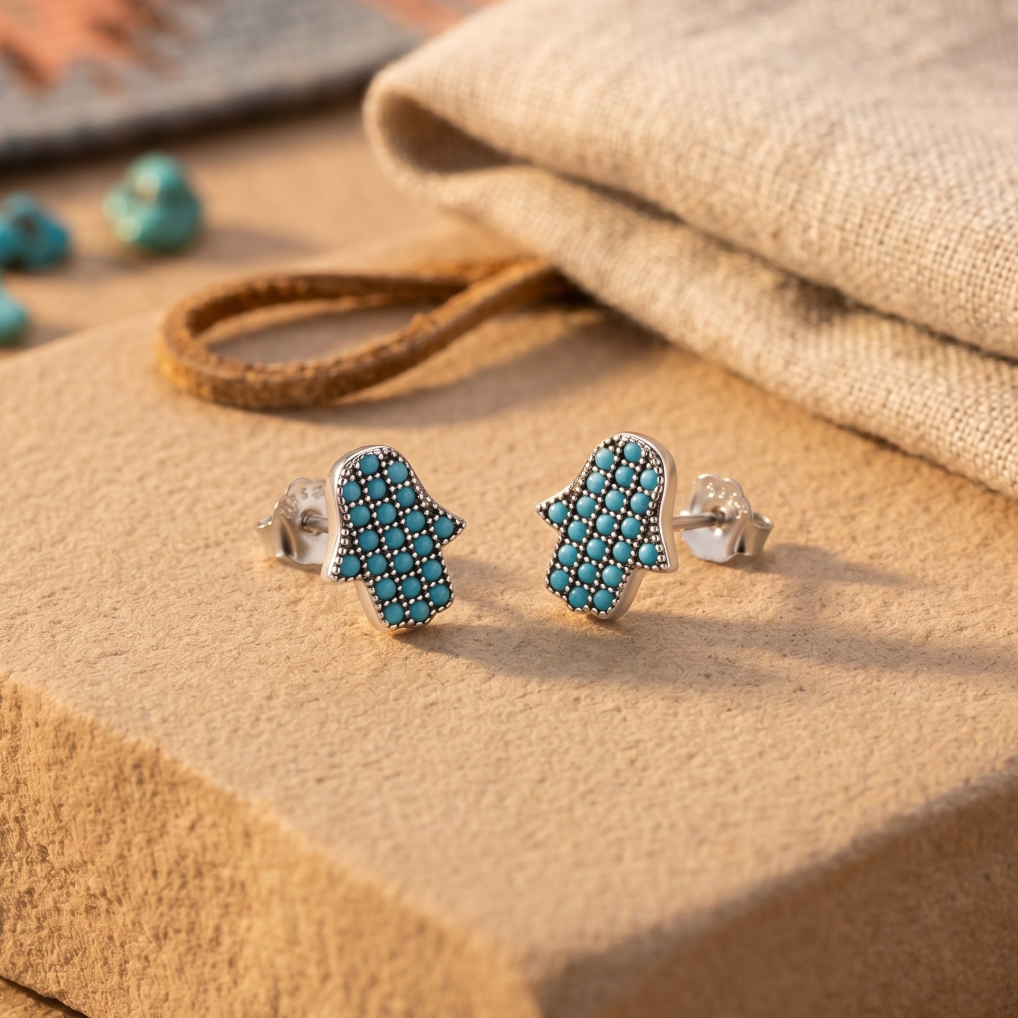 Silver Hamsa hand stud earrings with turquoise stones on a desert stone background.