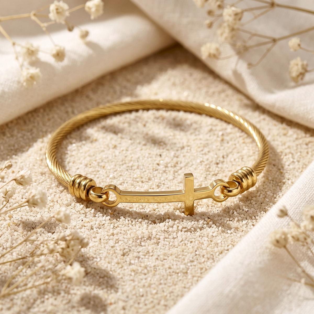 Gold-tone twisted cable bracelet with a horizontal cross, resting on pale sand among dried white flowers and fabric.