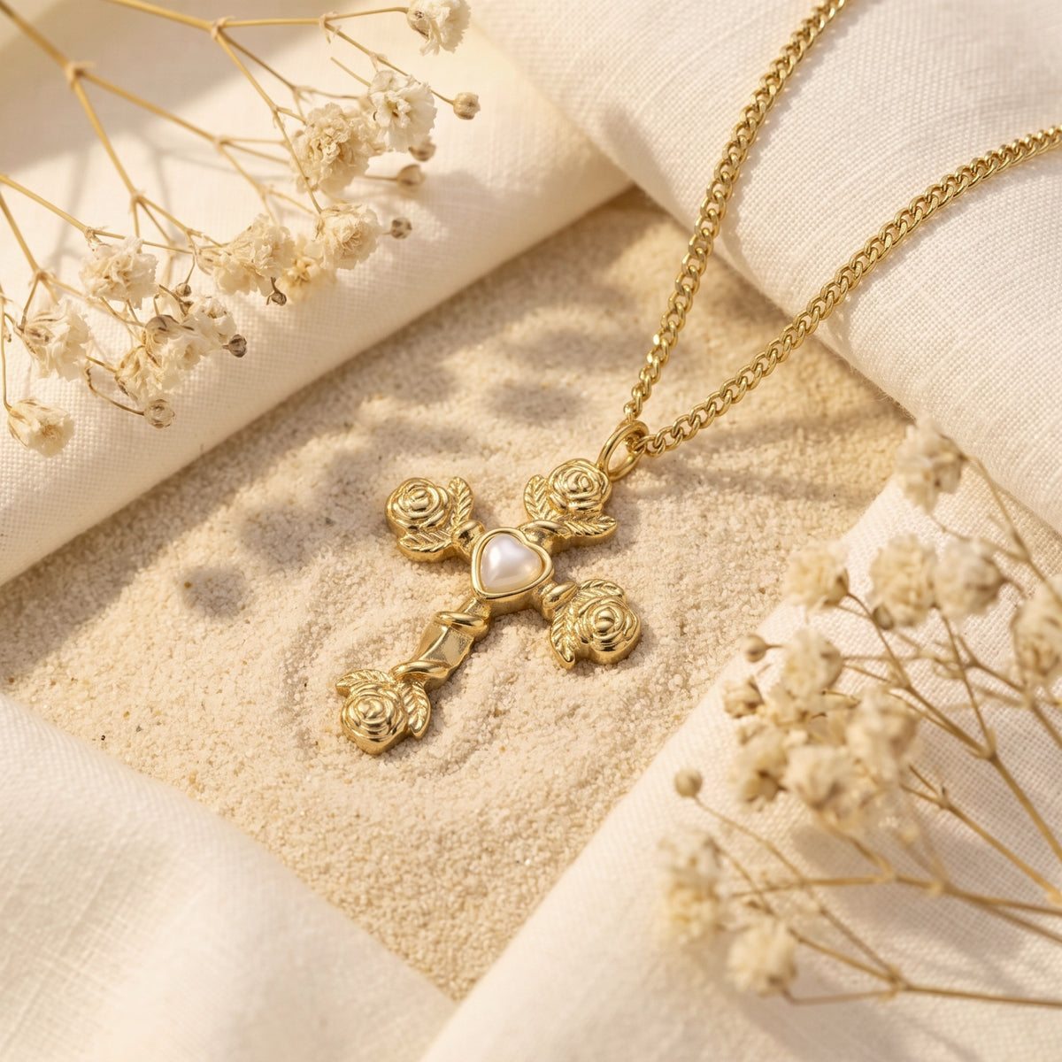Gold-tone rose cross necklace with a heart center resting on sand, framed by white fabric and dried flowers.